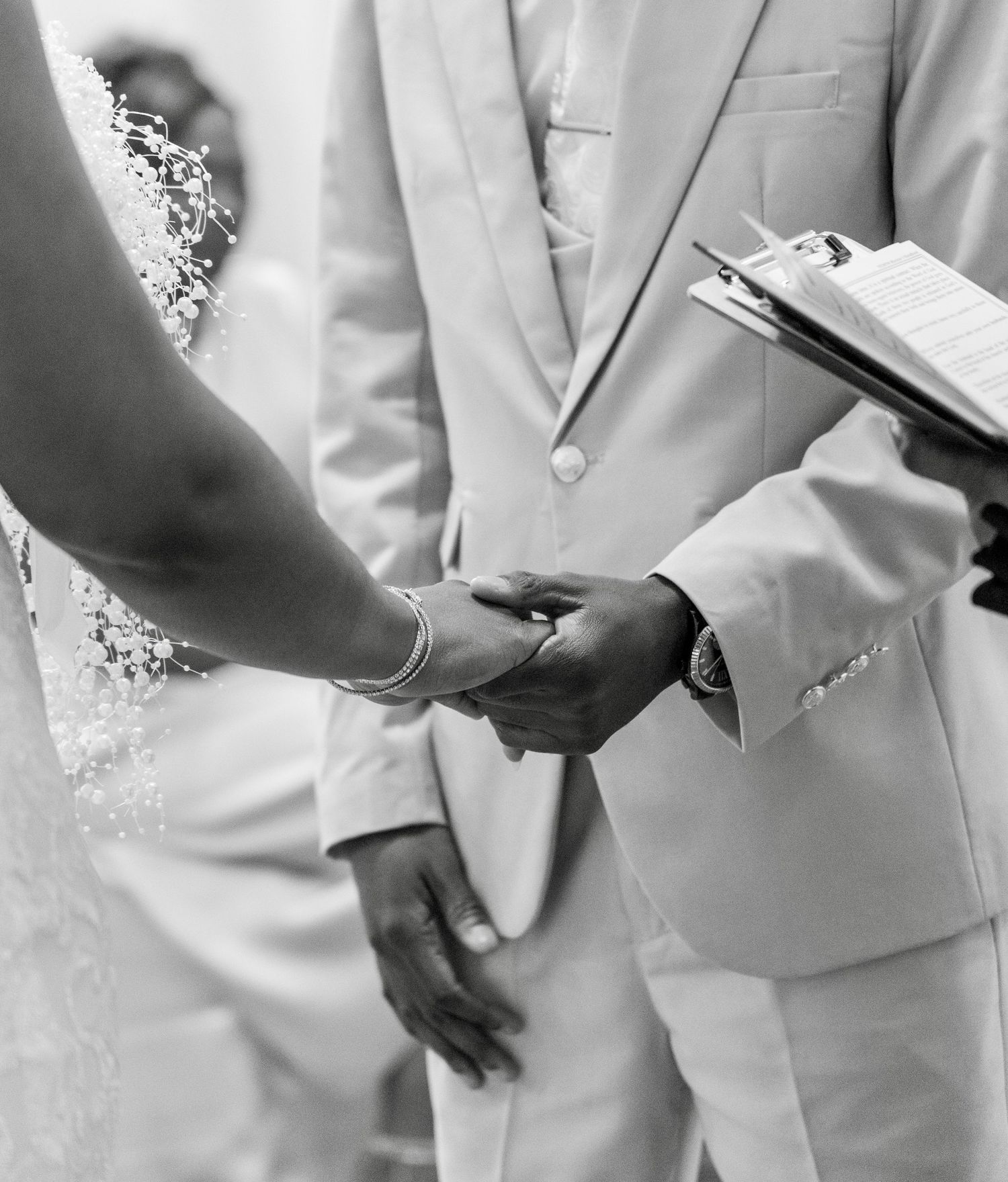 Black and white photo of a couple holding hands during a wedding ceremony, groom in white suit.