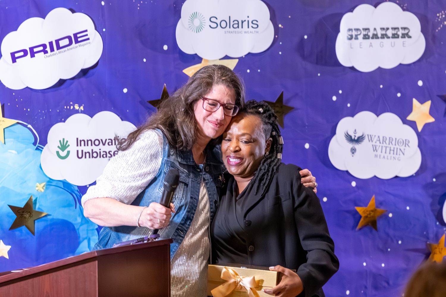 Two women sharing a warm hug on stage at a Solaris Pride event with sponsor banners in the background.