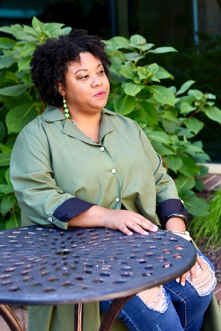 A Black woman in a green shirt and ripped jeans sits at an outdoor metal table surrounded by lush greenery.
