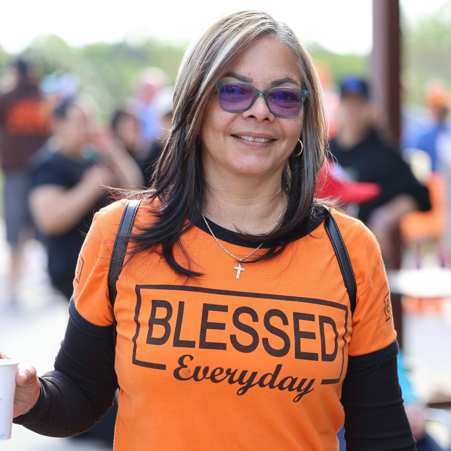 Woman smiling in orange Blessed Everyday t-shirt with purple sunglasses at outdoor community event. Multiple Sclerosis. Walk MS