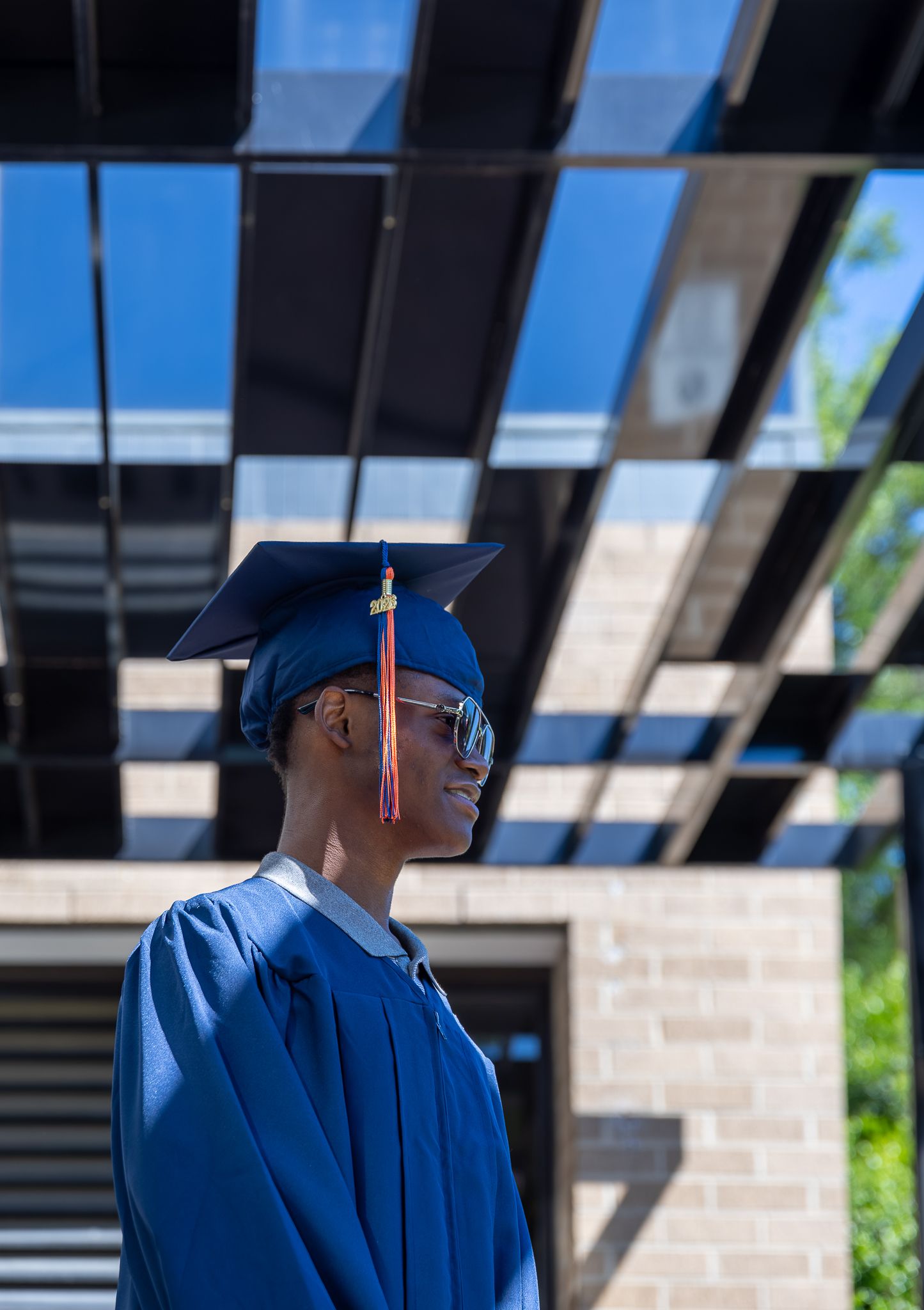 Graduate in blue cap and gown smiling outdoors under modern pergola on a sunny graduation day.
