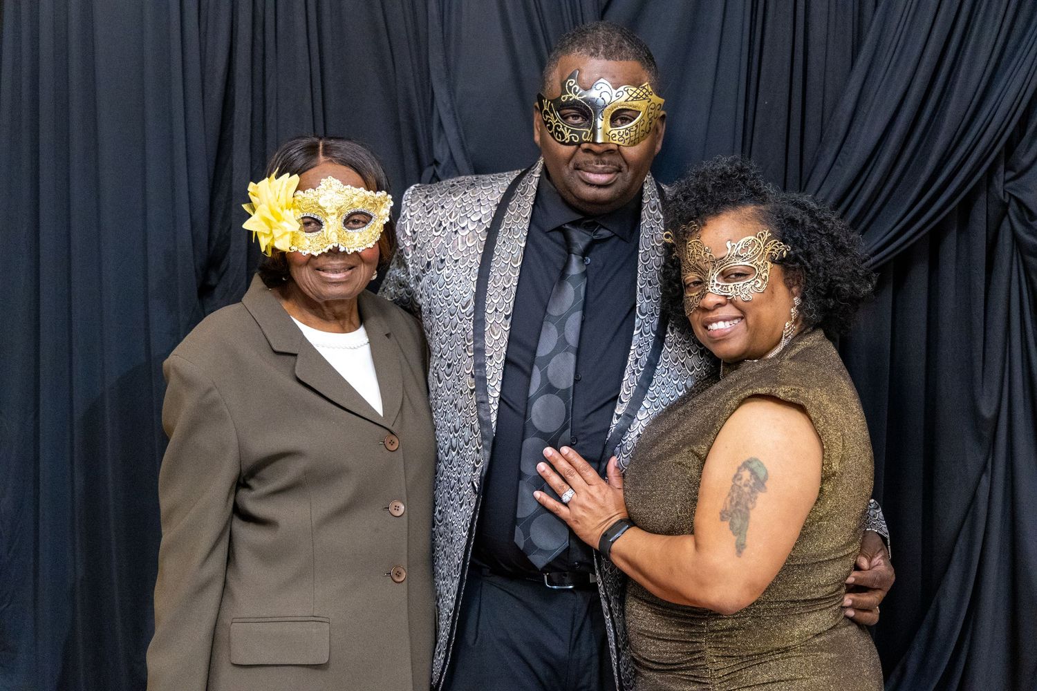 Three people wearing gold masquerade masks posing together against black drape backdrop at formal event.