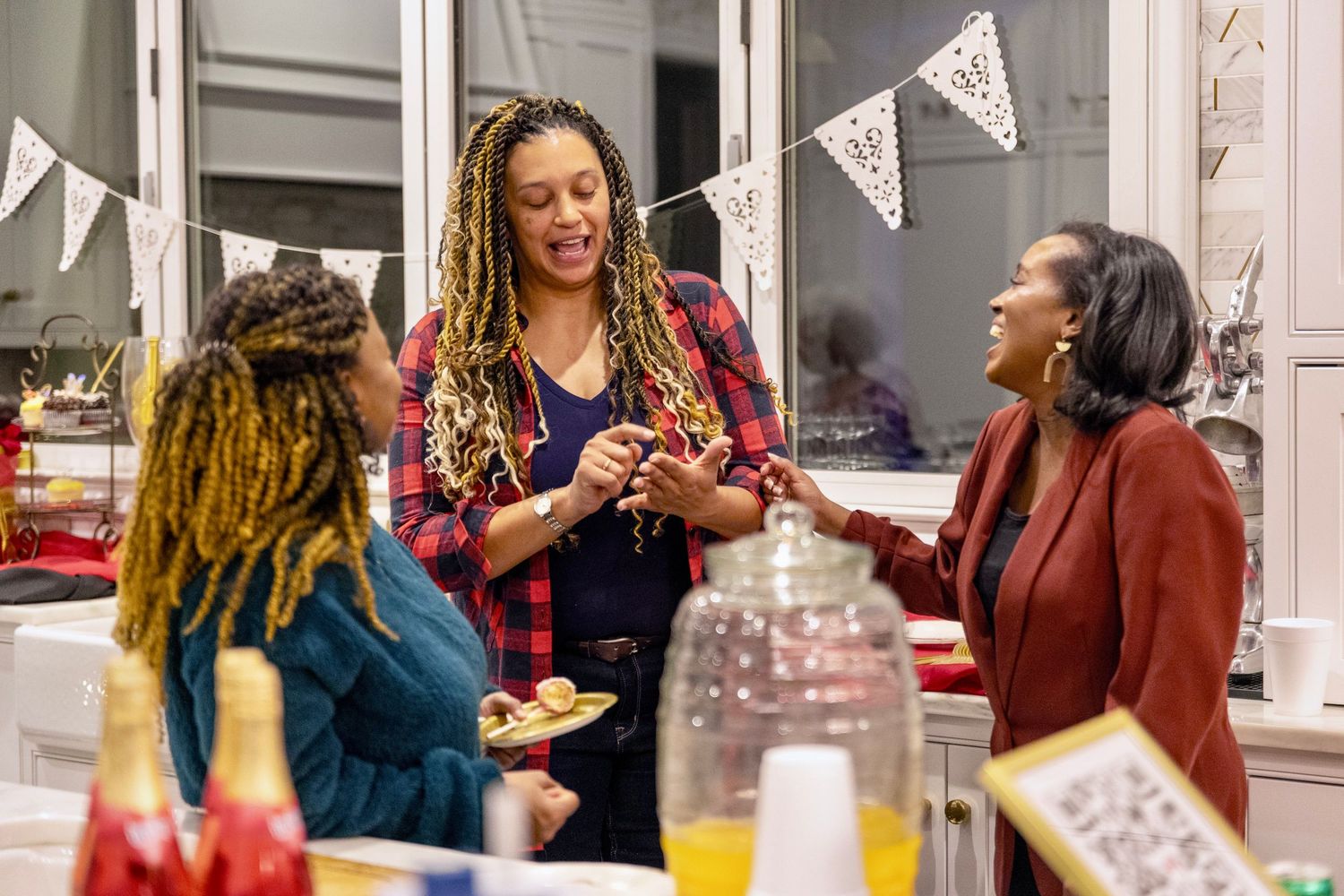 Three women laughing and socializing at indoor celebration party with bunting decorations and drinks on table. Bride