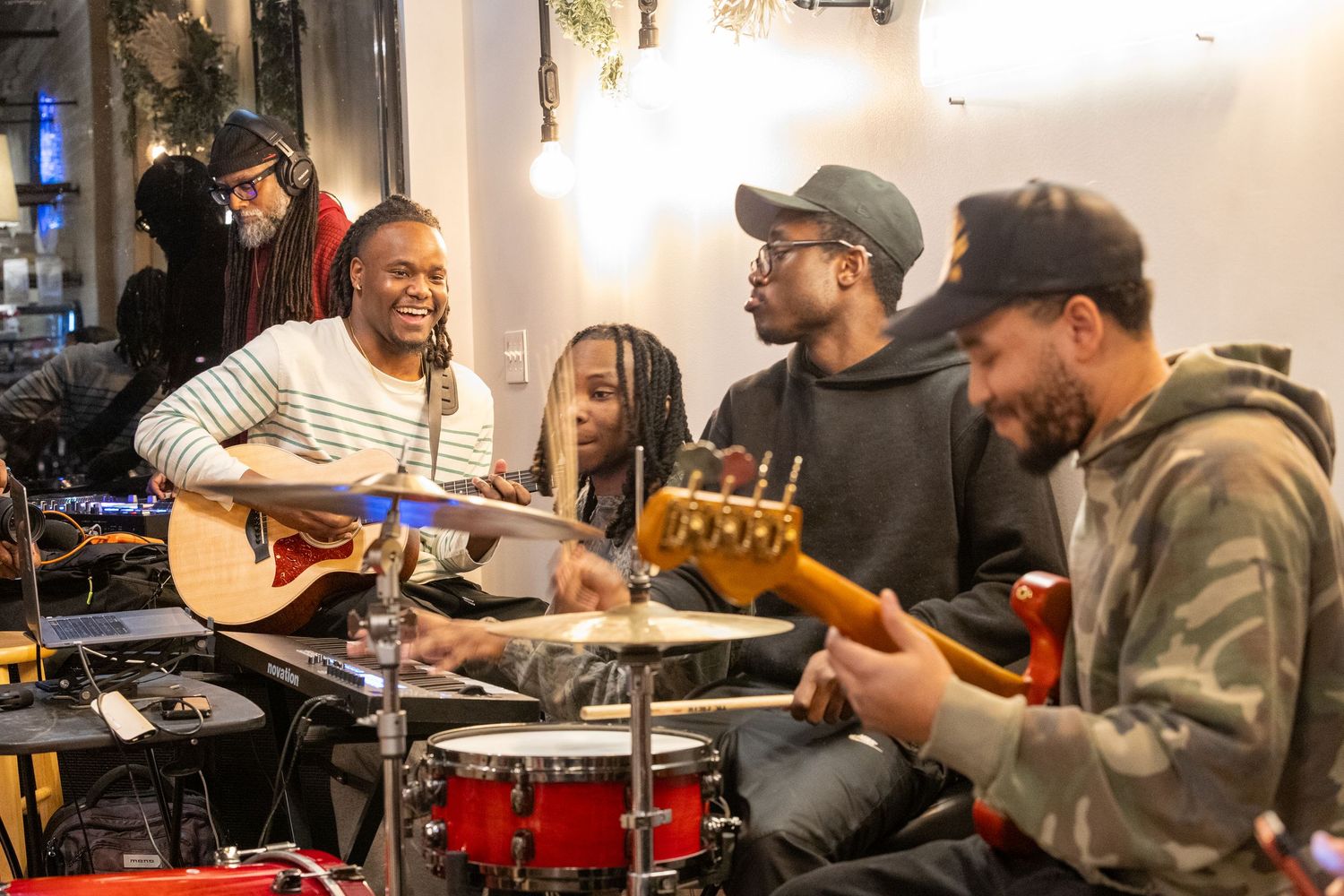 A smiling musician performs with bandmates playing guitar, drums, and bass in a cozy venue.