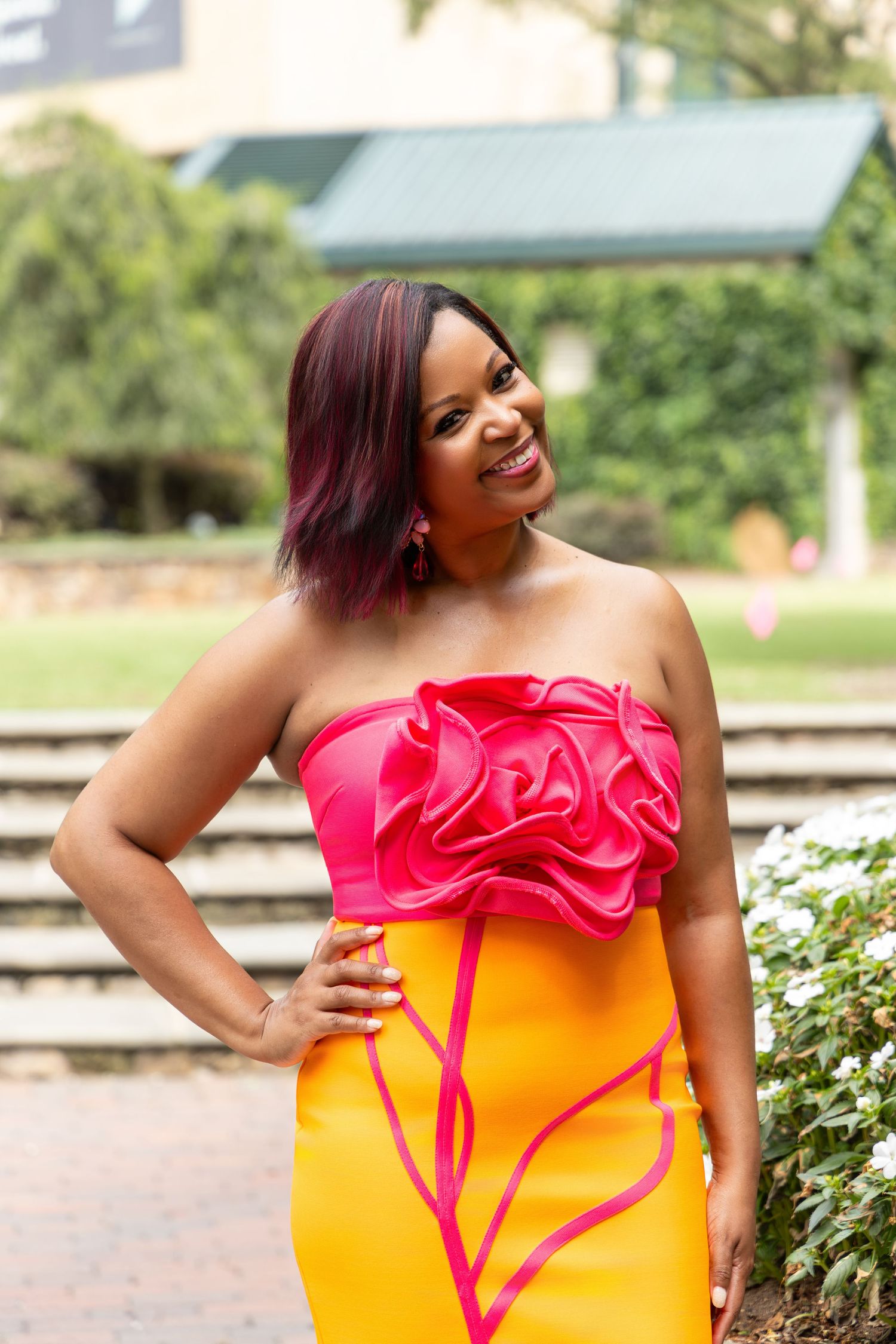Woman in pink strapless floral top and yellow skirt smiling outdoors in a garden setting.
