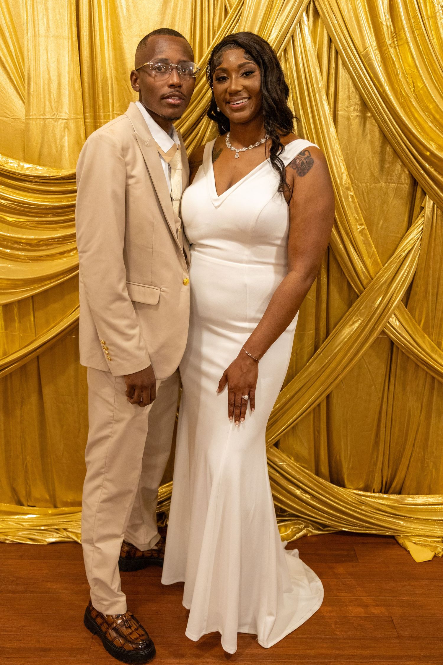African American bride and groom posing together in front of gold drape backdrop at wedding.