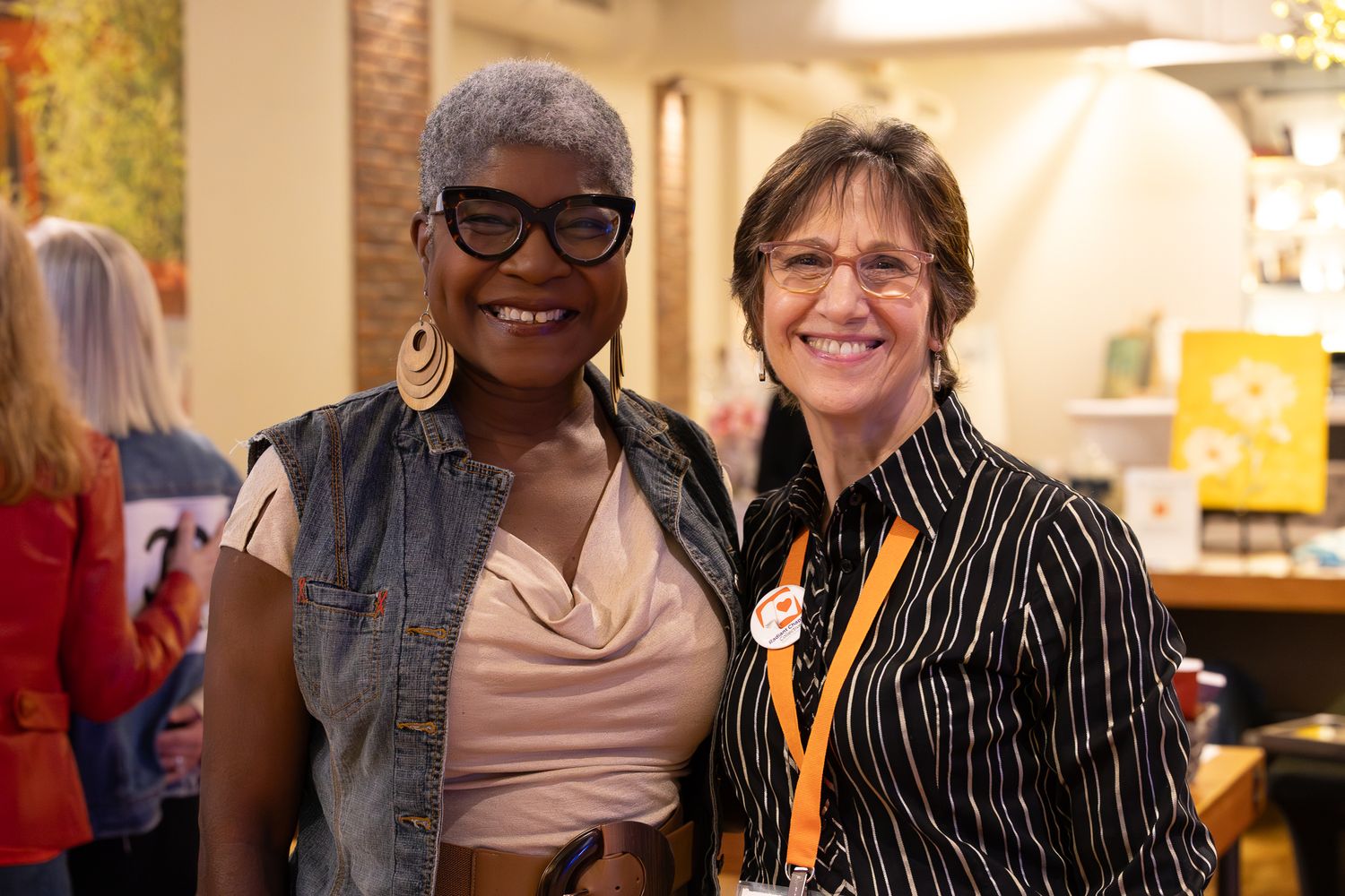 Two smiling women pose together at an indoor event, one wearing glasses and hoop earrings, the other in a striped shirt.