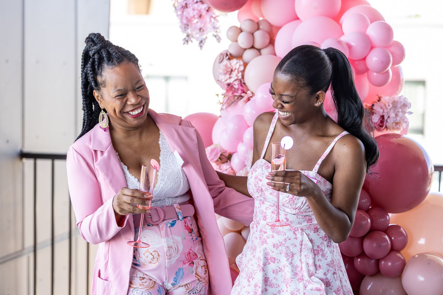 Two Black women laugh together at a pink baby shower, holding champagne glasses in front of a pink balloon arch.