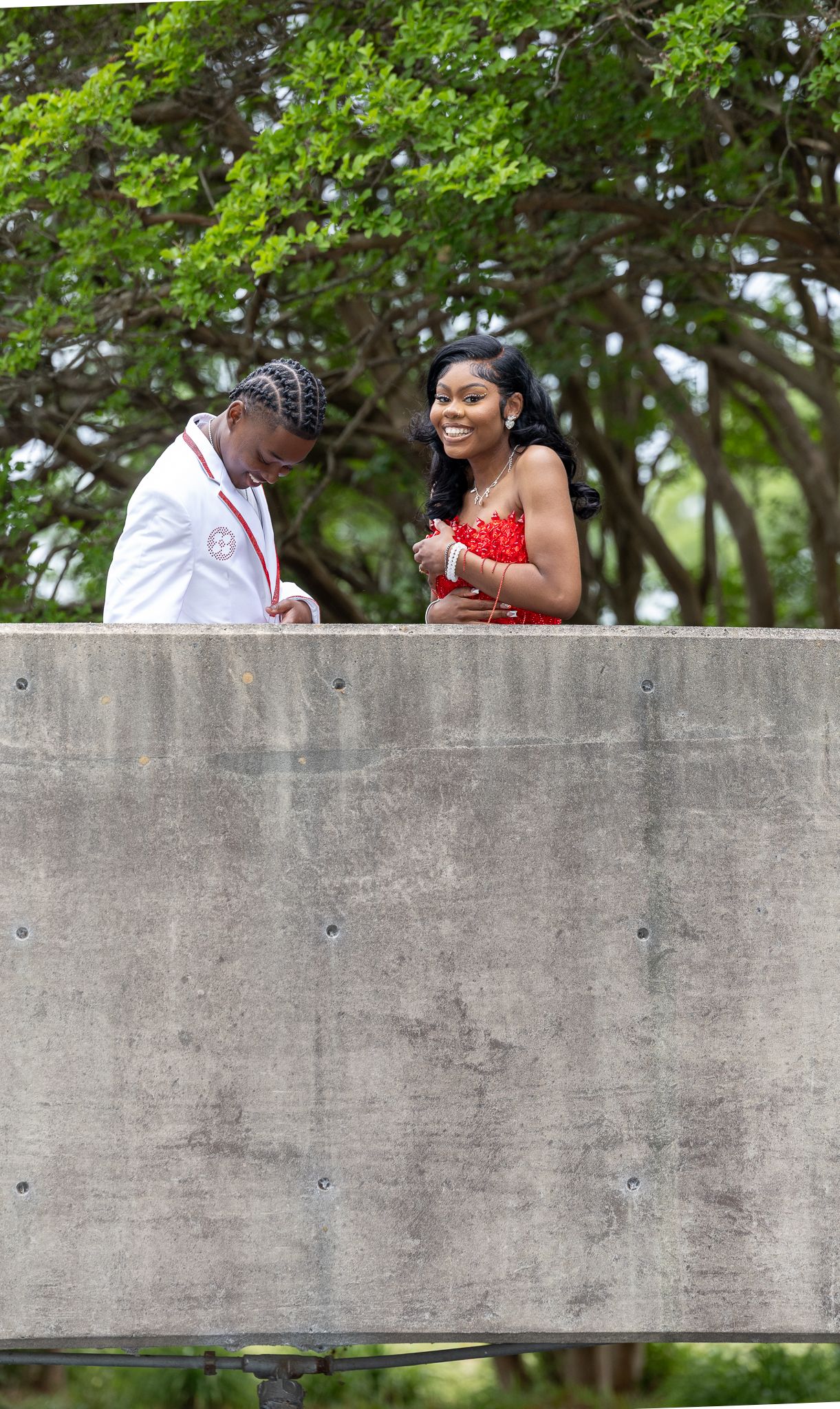 A smiling couple in formal attire stands on an elevated platform outdoors, surrounded by lush green trees. Prom