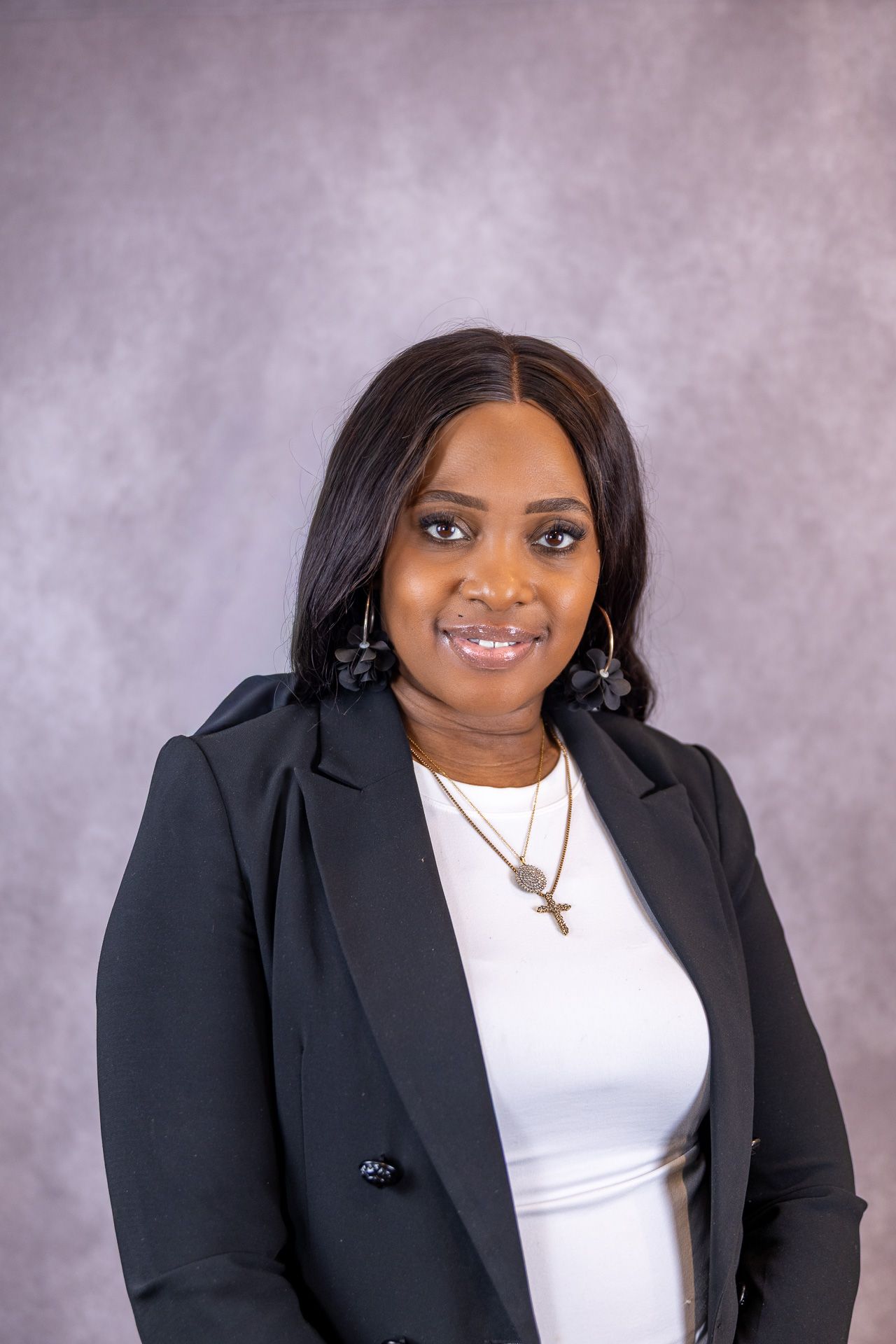 A confident Black woman in a black blazer smiles in a professional headshot with hoop earrings and gold necklace.