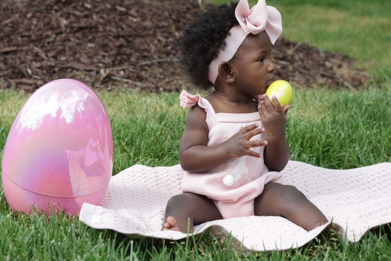An adorable Black baby girl in pink romper sits on a blanket outdoors, chewing a yellow Easter egg beside a large pink egg.
