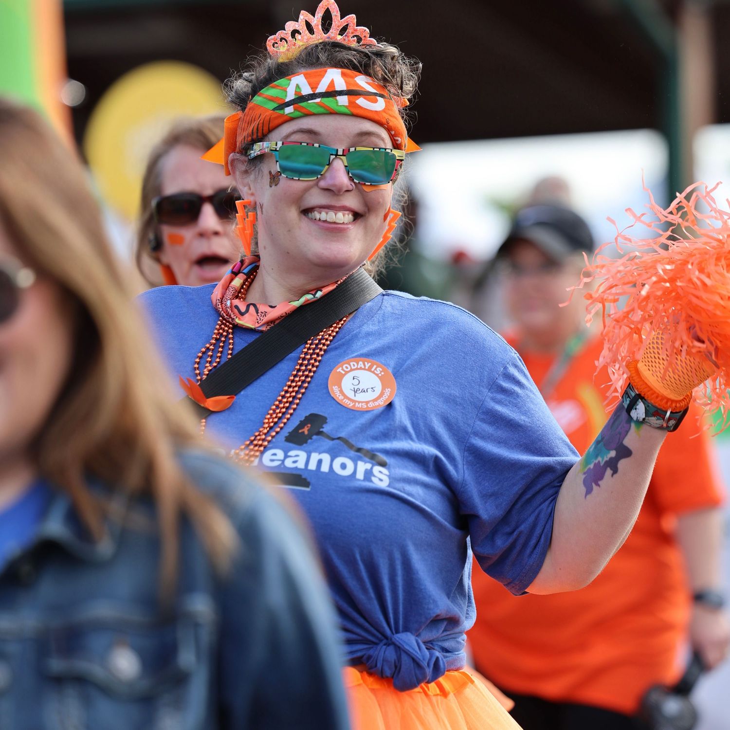 Woman in orange MS Walk gear smiling at charity event wearing festive accessories and blue team shirt.