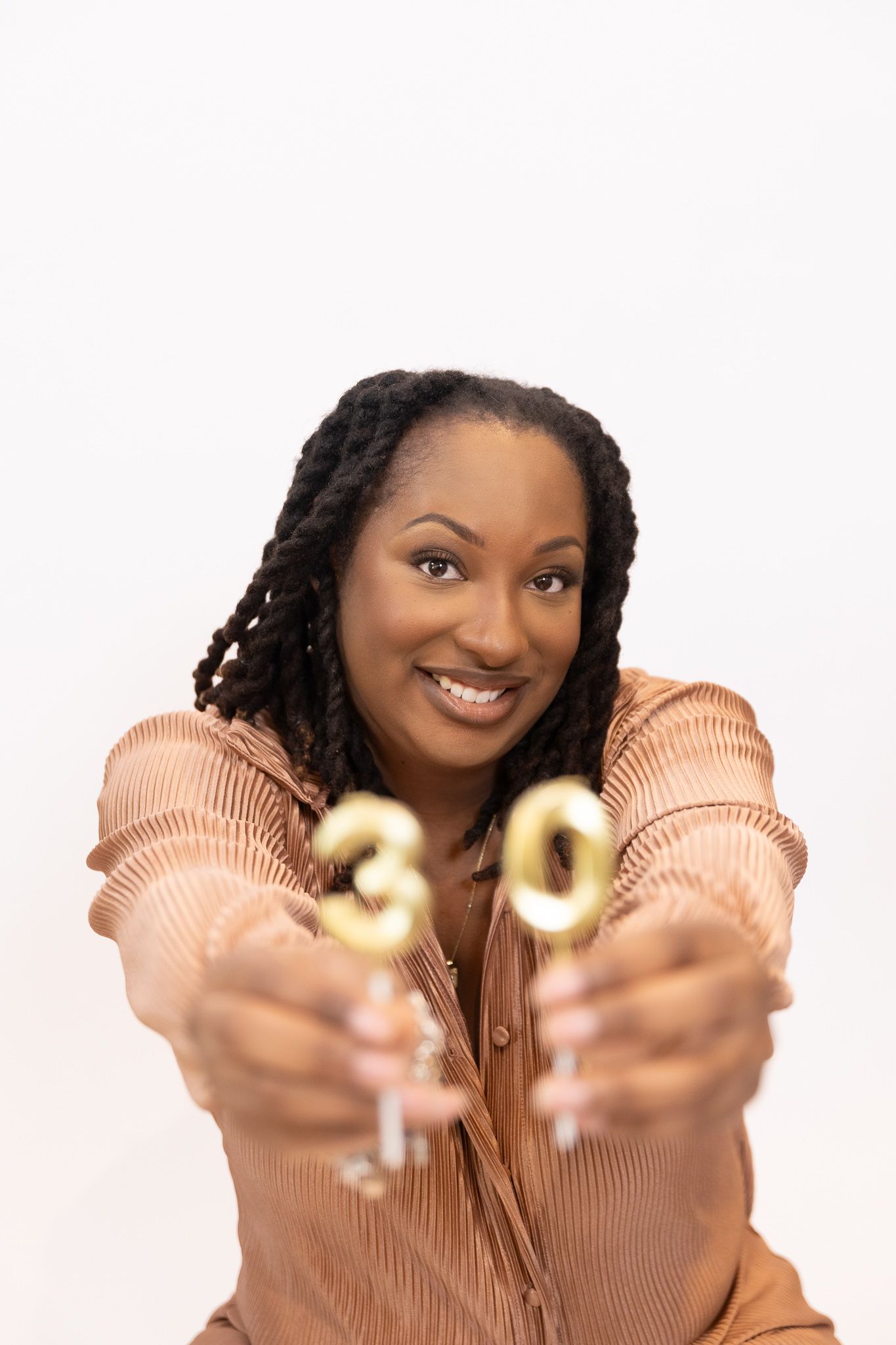 Smiling Black woman with braids holds gold number 30 candles toward camera on white background.