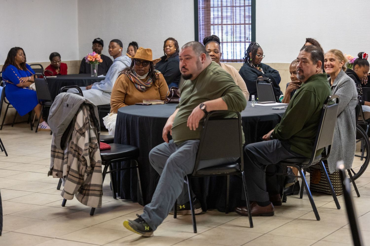 Diverse group of adults seated at round tables during a community meeting or workshop in a hall.