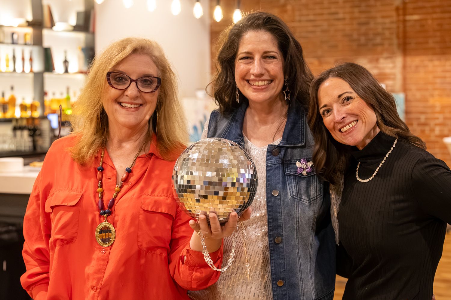 Three women smiling and posing together at a bar, with the middle woman holding a shiny disco ball.