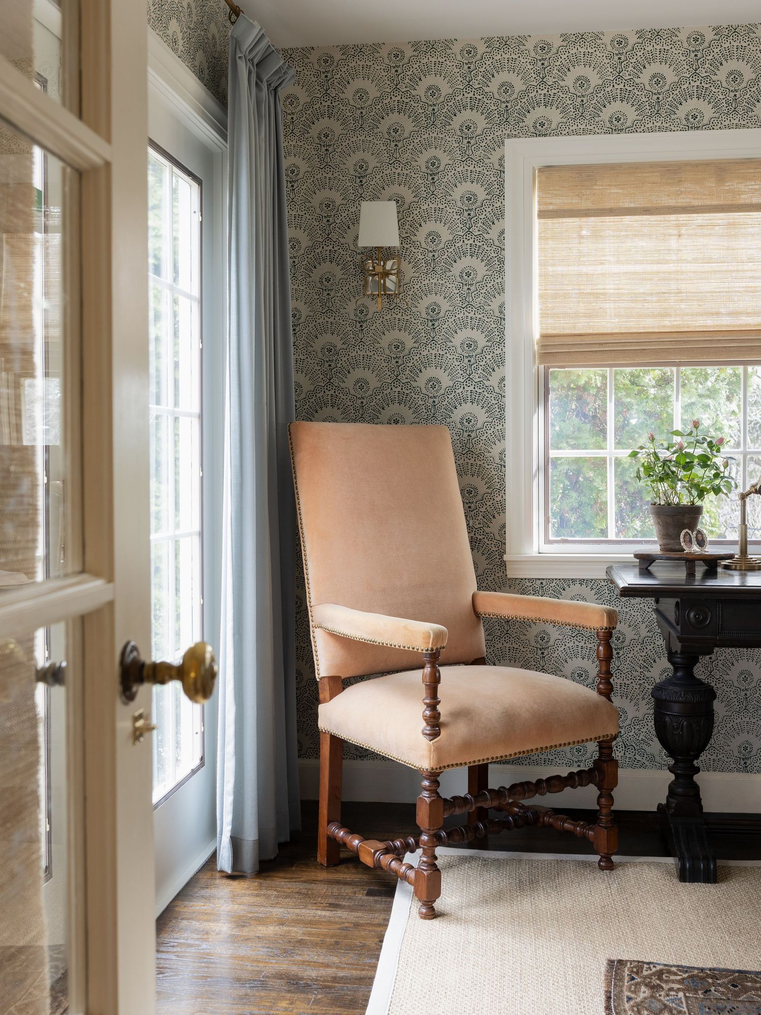 Elegant room with patterned wallpaper, antique upholstered chair, hardwood floors, Roman shade, and French door with blue curtains.