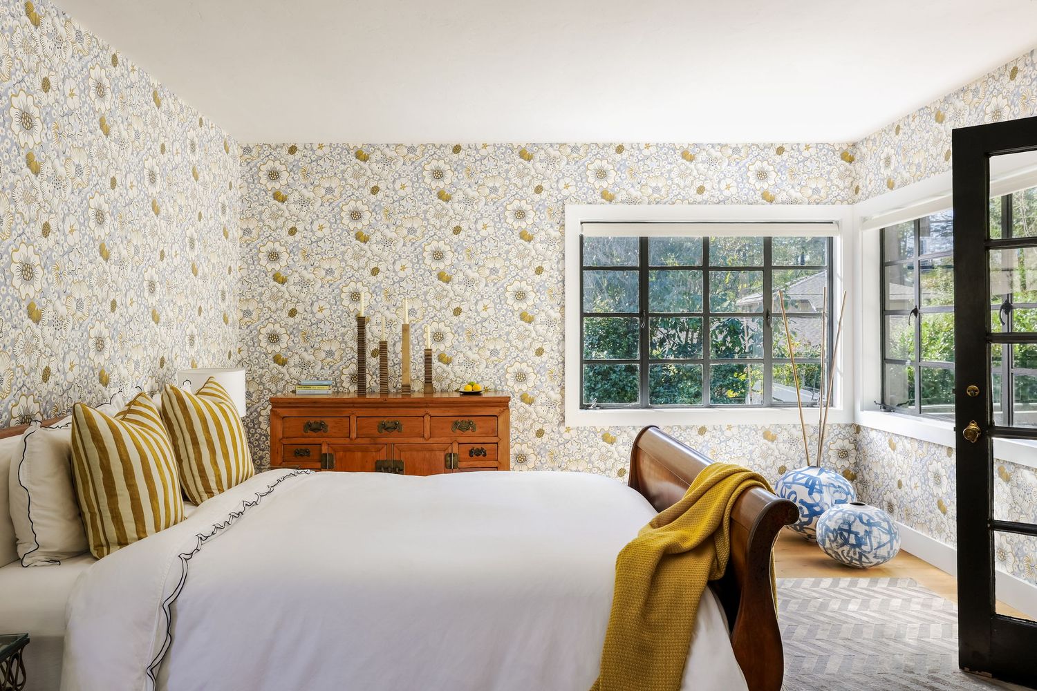 Floral wallpaper bedroom with white bedding, antique dresser, striped pillows, yellow throw, and garden window view.