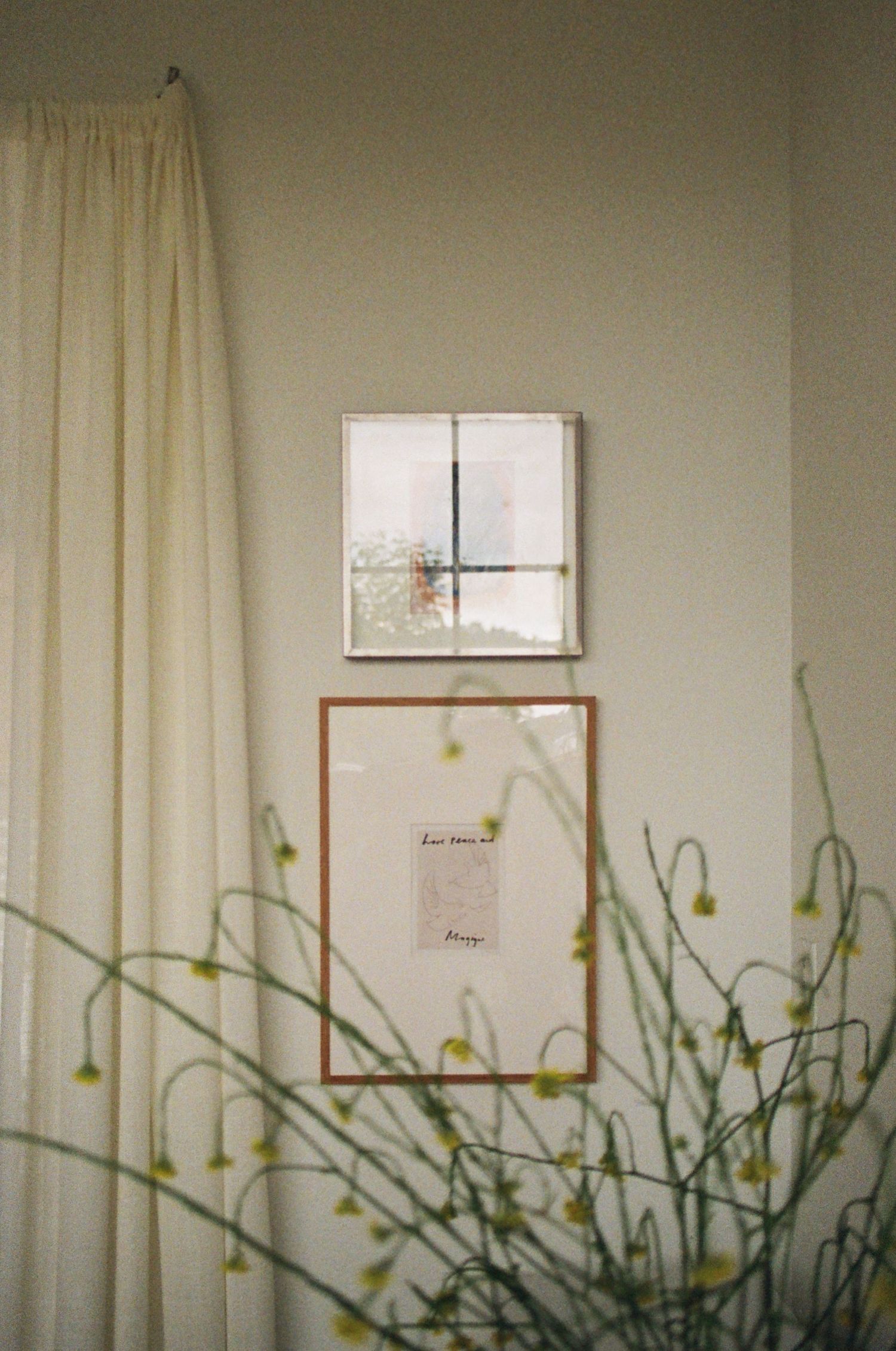A cozy room with a window, curtains, and a framed artwork on the wall, surrounded by potted plants.