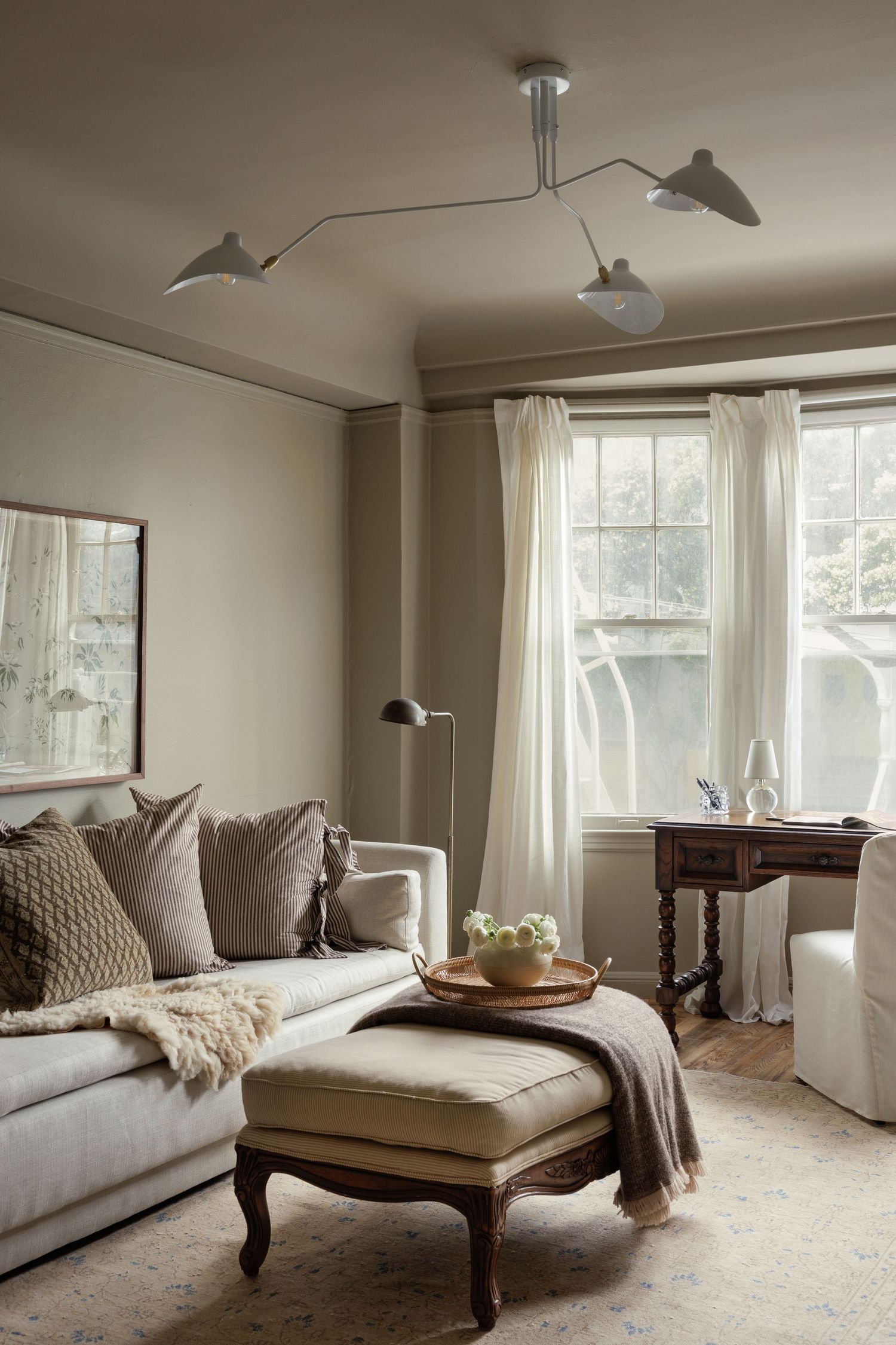 Cozy neutral living room with a linen sofa, ottoman, Serge Mouille-style ceiling light, and sheer curtained windows.
