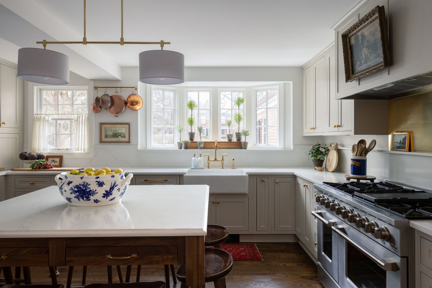 Elegant white kitchen with brass pendant lights, farmhouse sink, stainless range, and island with blue bowl centerpiece.