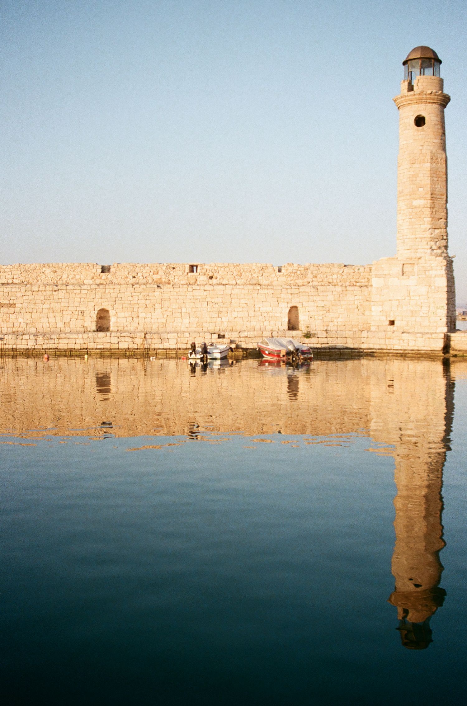 Historic lighthouse and stone fortress wall reflecting in calm harbor waters.