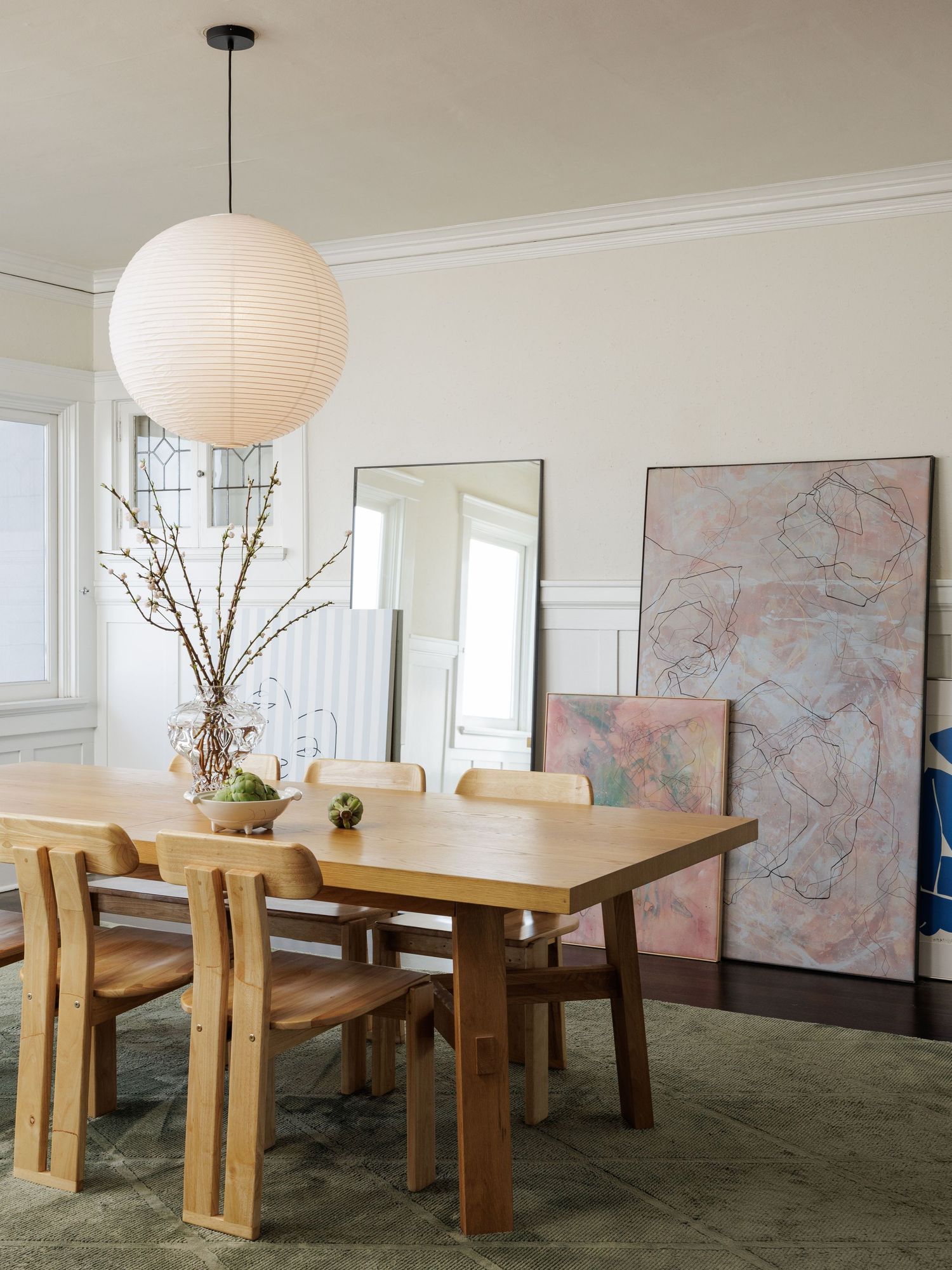 Modern dining room with wooden table, chairs and paper lantern pendant light against white walls with pink artwork.
