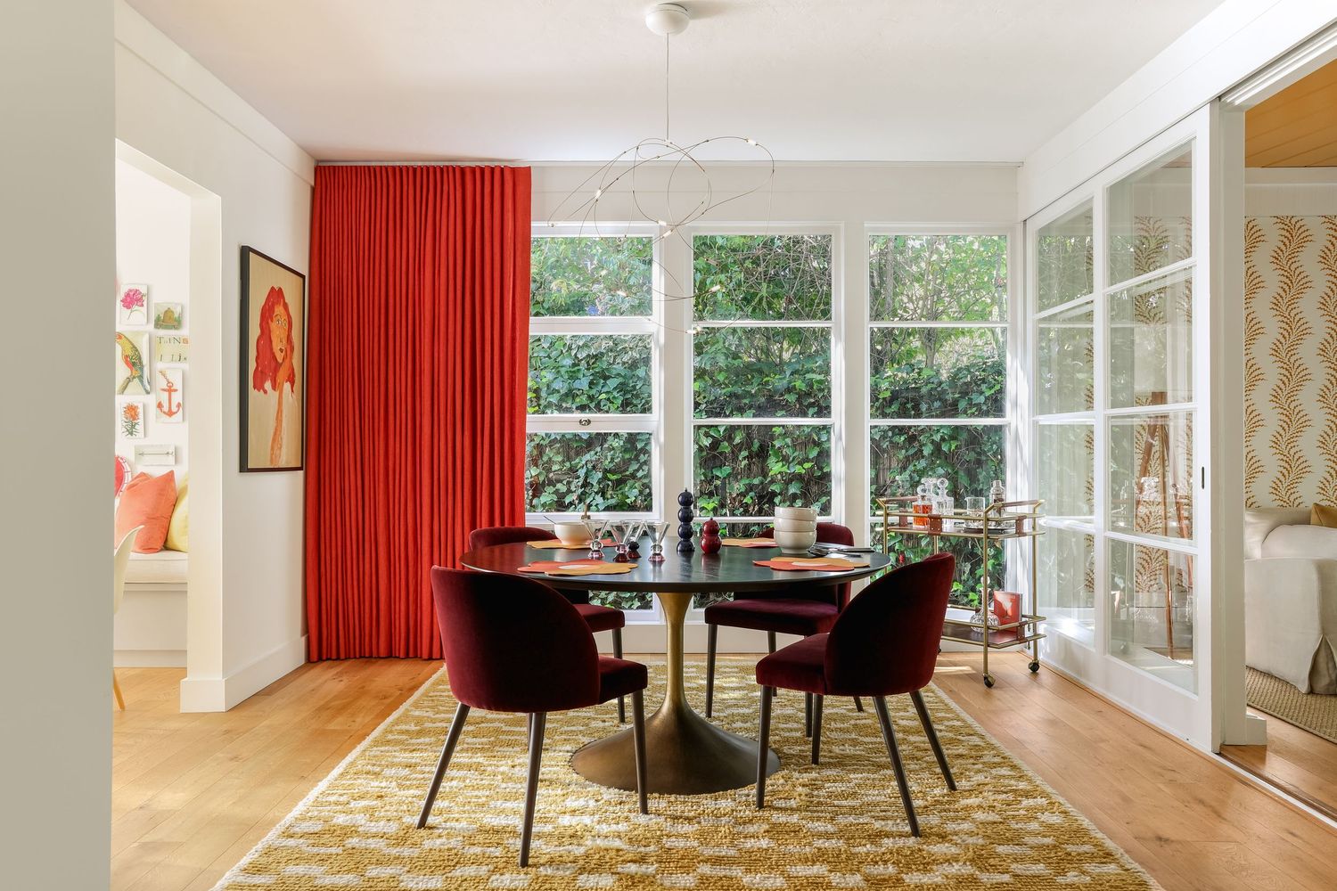 Bright dining room with red curtains, tulip table, velvet chairs, patterned rug, and large windows overlooking greenery.