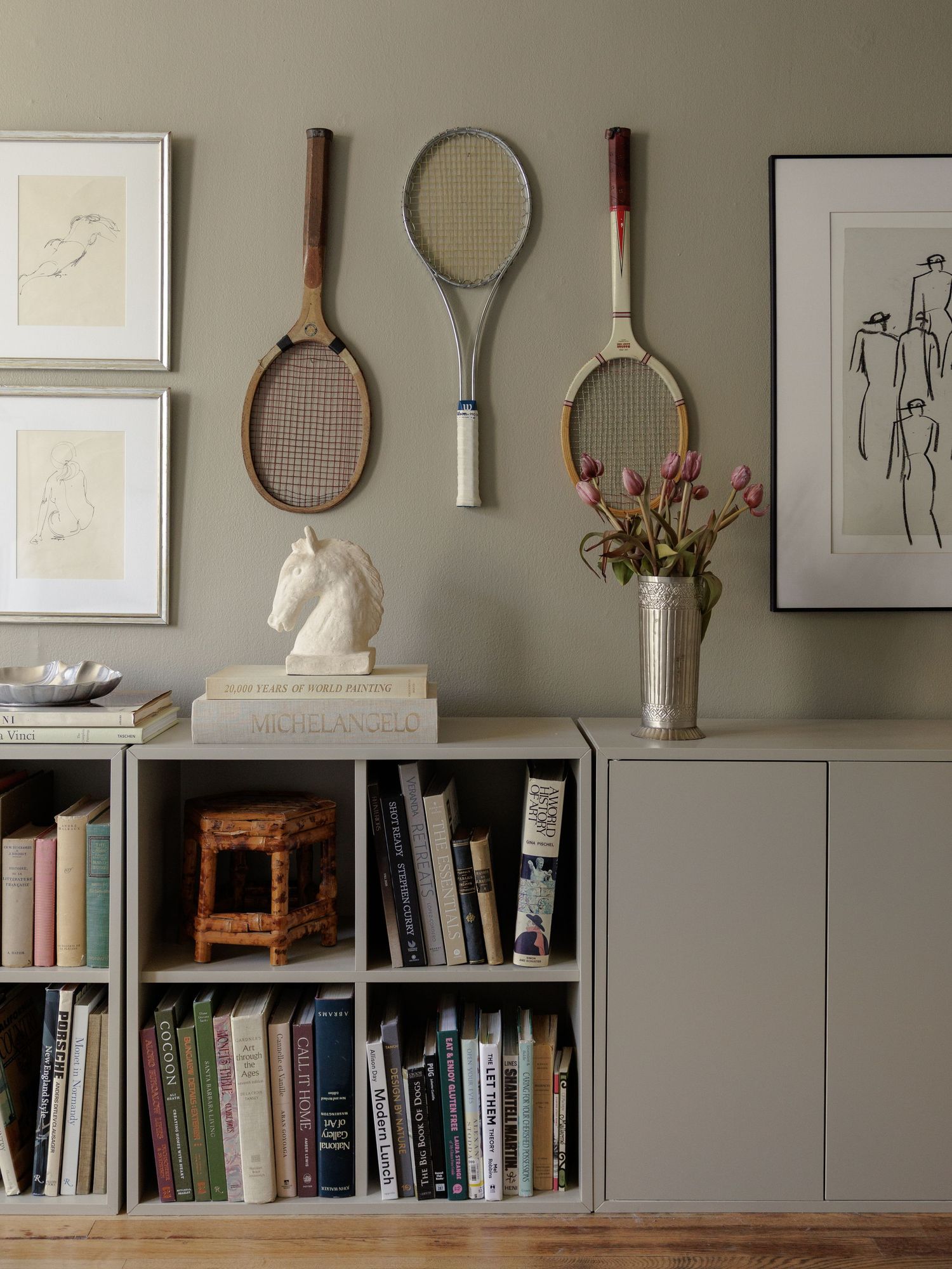 Vintage tennis rackets displayed on sage green wall above gray bookcase filled with books, beside framed art and floral arrangement.