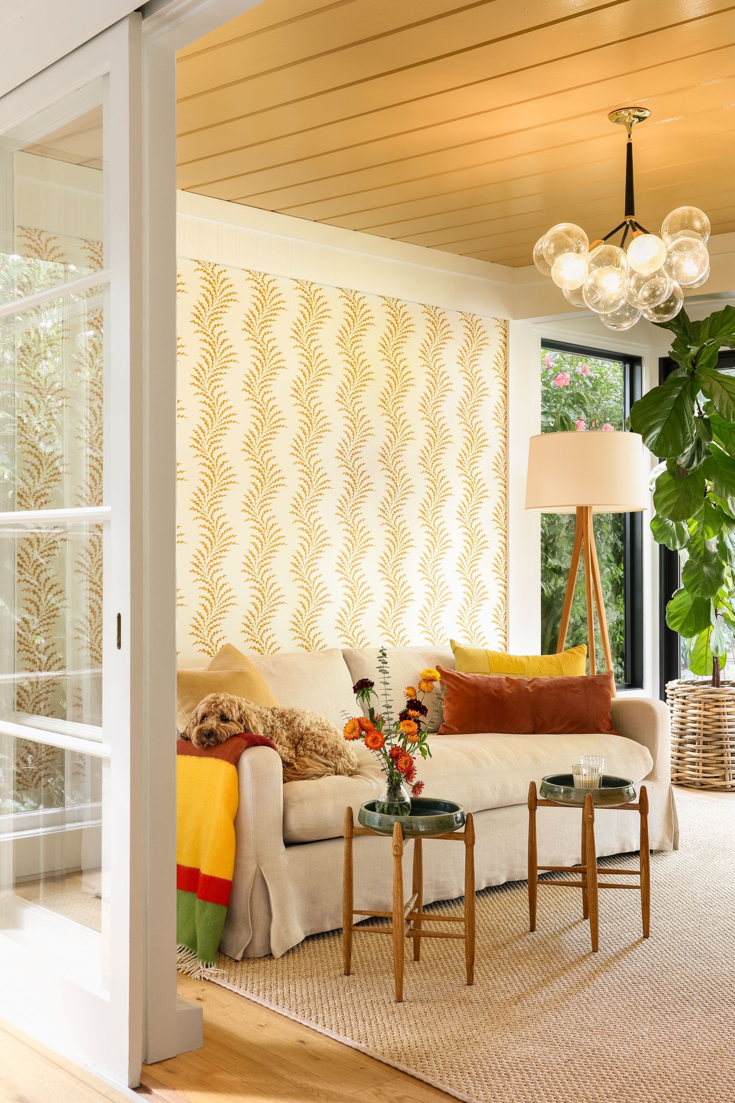 Bright living room with gold leaf wallpaper, cream sofa, colorful pillows, wood ceiling, globe chandelier, and tropical plants.