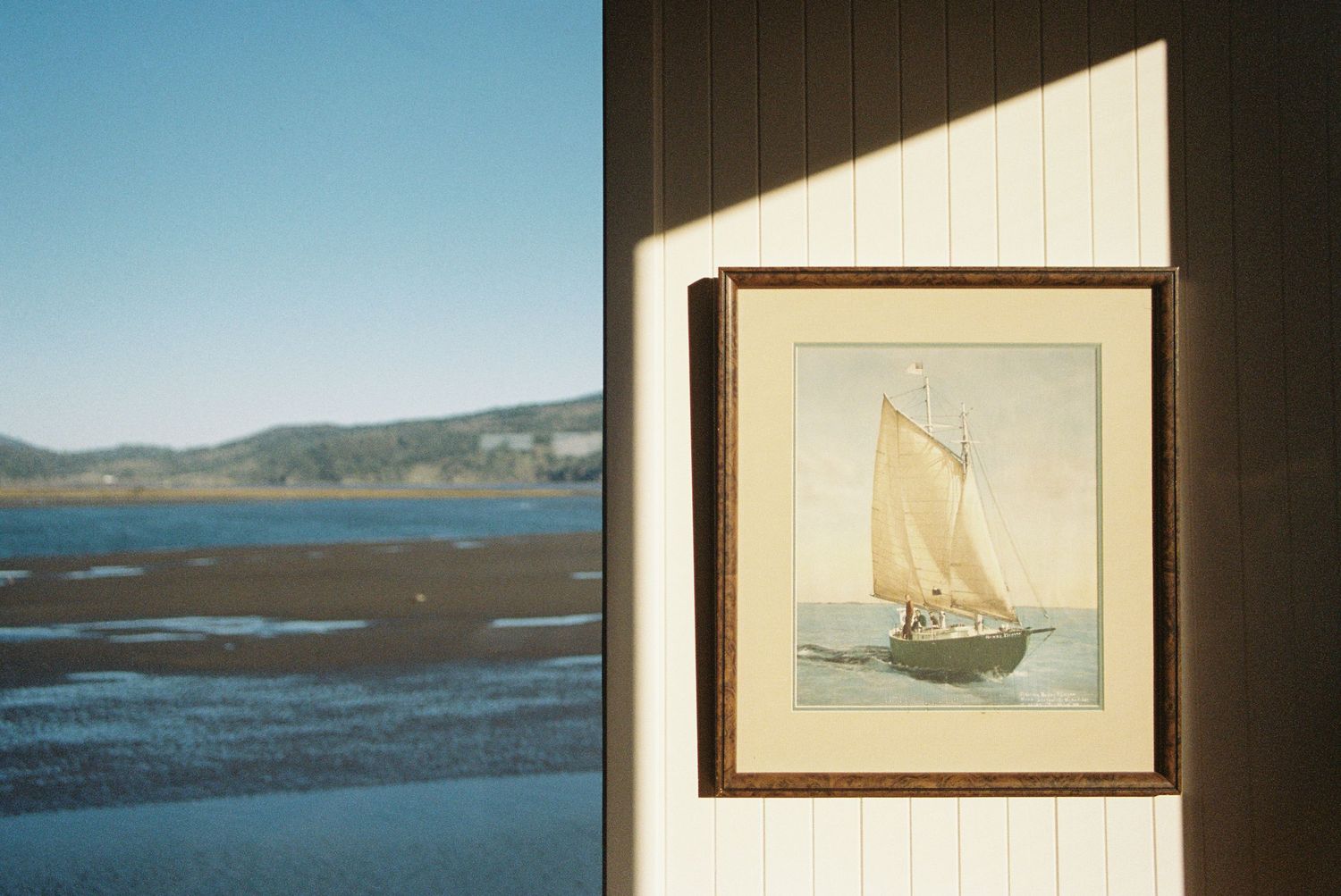 Framed sailboat painting on white wall beside coastal water view under blue sky.