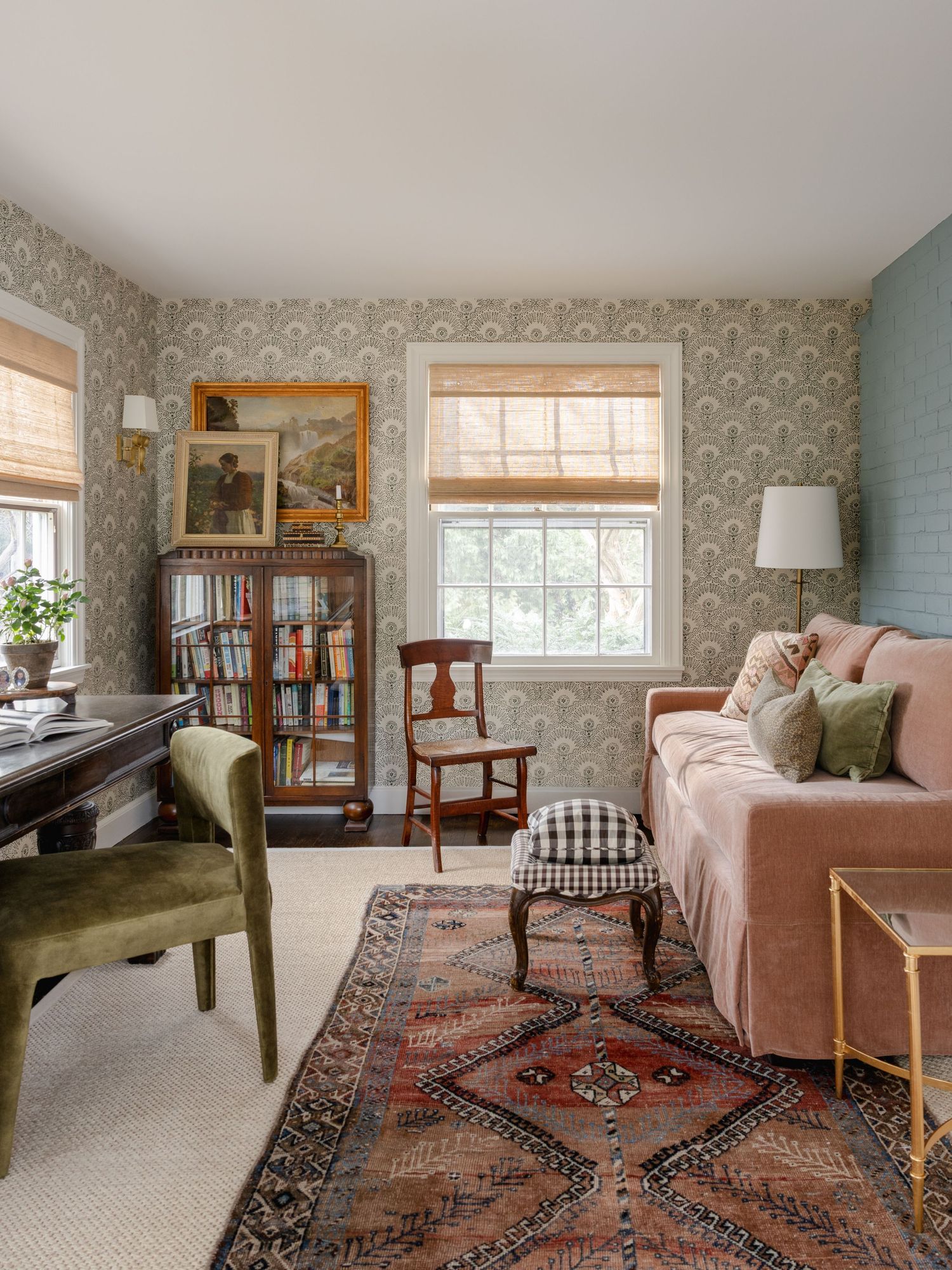 Cozy home office with pink velvet sofa, Persian rug, patterned wallpaper, wooden bookcase, and green velvet chair near a sunlit window.