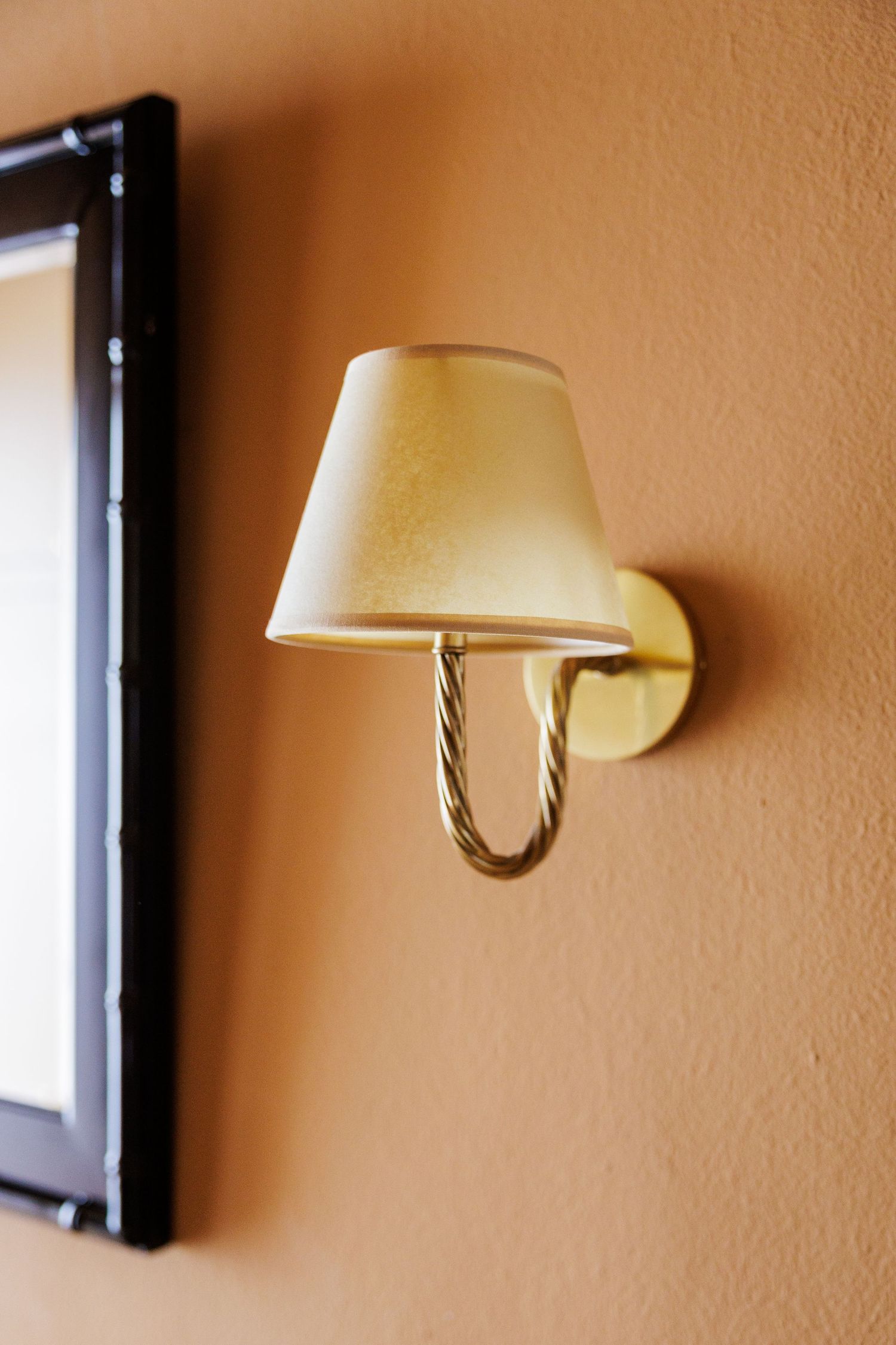 Elegant wall sconce with cream lampshade and twisted brass arm mounted on warm terracotta wall beside framed artwork.