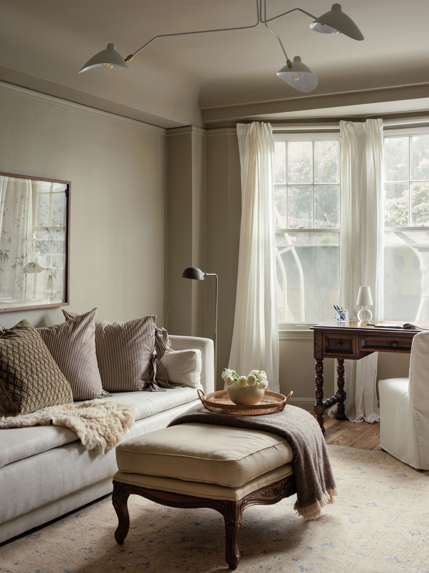 Cozy neutral living room with beige sofa, tufted ottoman, bay window, and mid-century sputnik ceiling light.