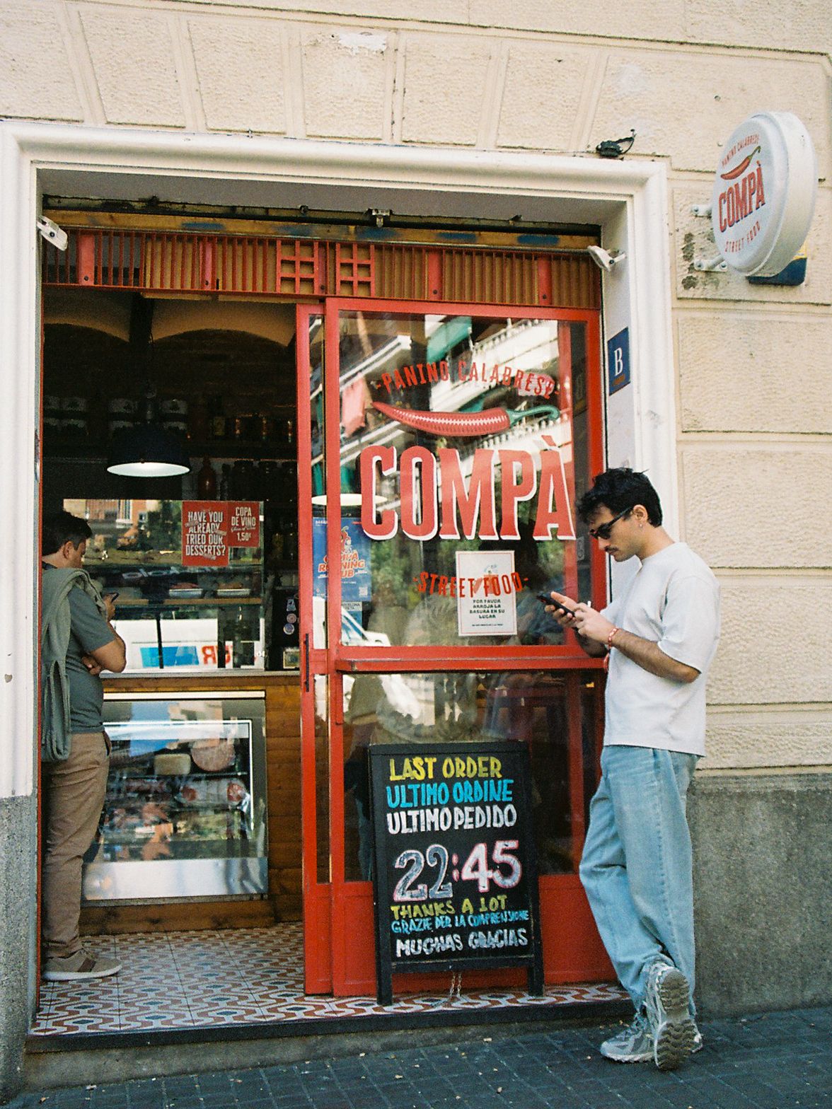 Man checks phone outside Compa sandwich shop with red storefront and last order sign at 22:45.