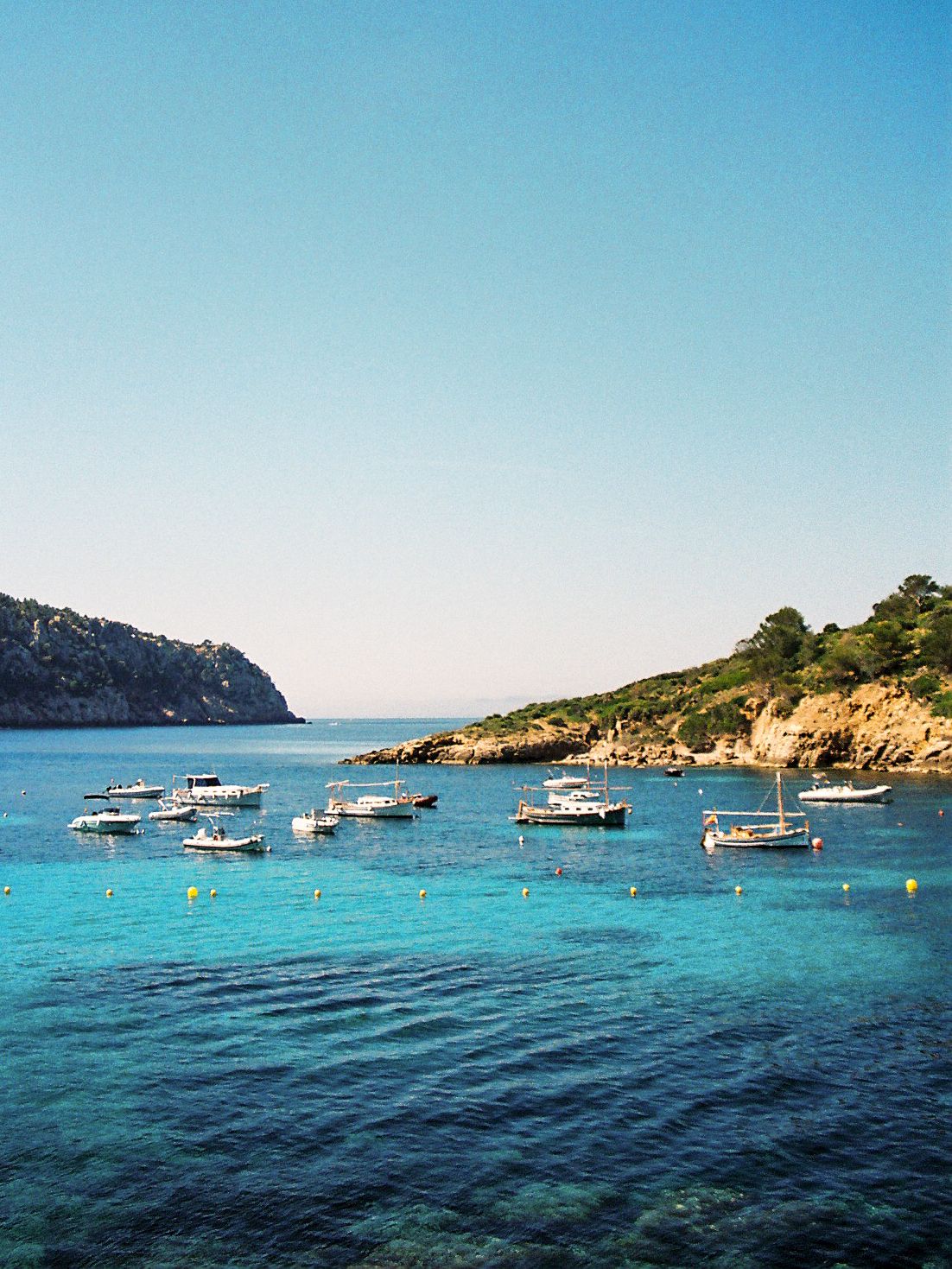 Several small boats anchored in a stunning turquoise bay surrounded by rocky hills under a clear blue sky.