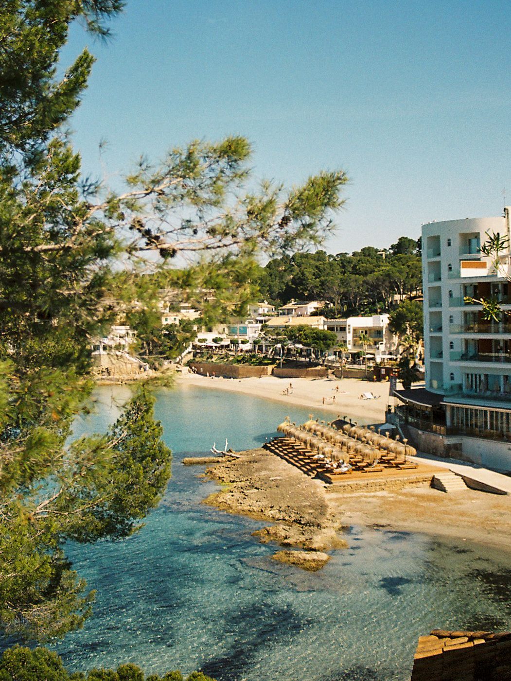 Scenic coastal cove in Ibiza with crystal-clear turquoise water, sandy beach, sun loungers, and white Mediterranean buildings.
