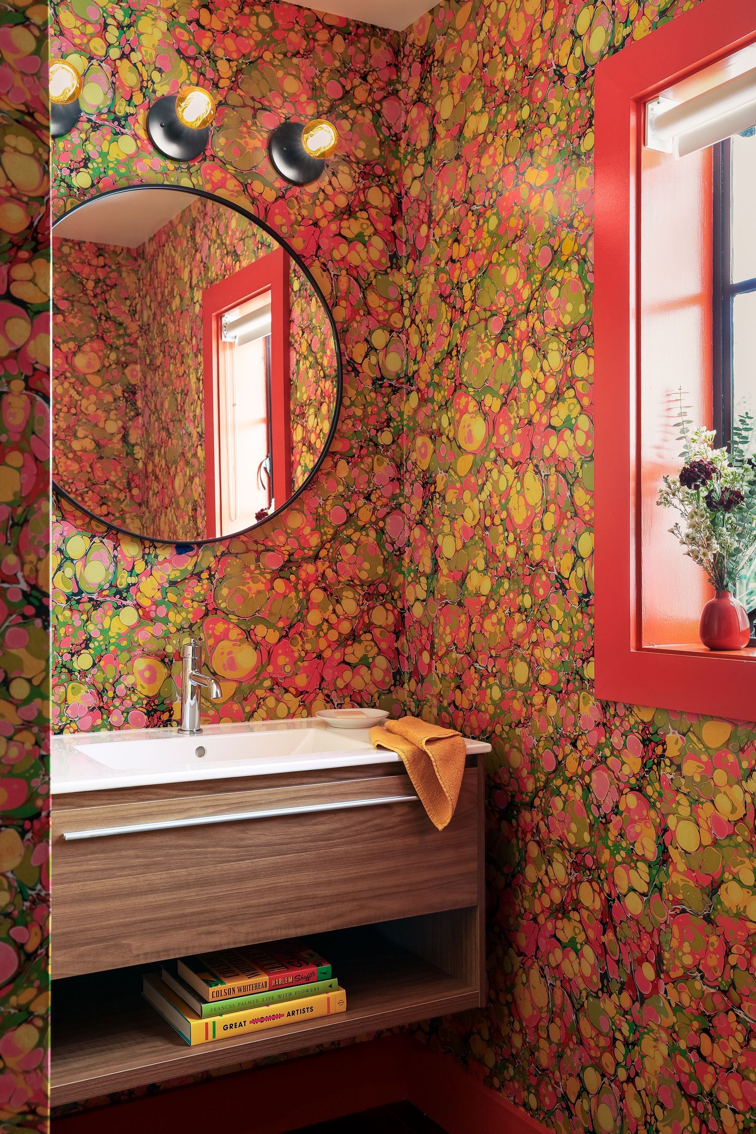 Vibrant marbled wallpaper bathroom with round mirror, floating wood vanity, red window trim, and colorful books on shelf.