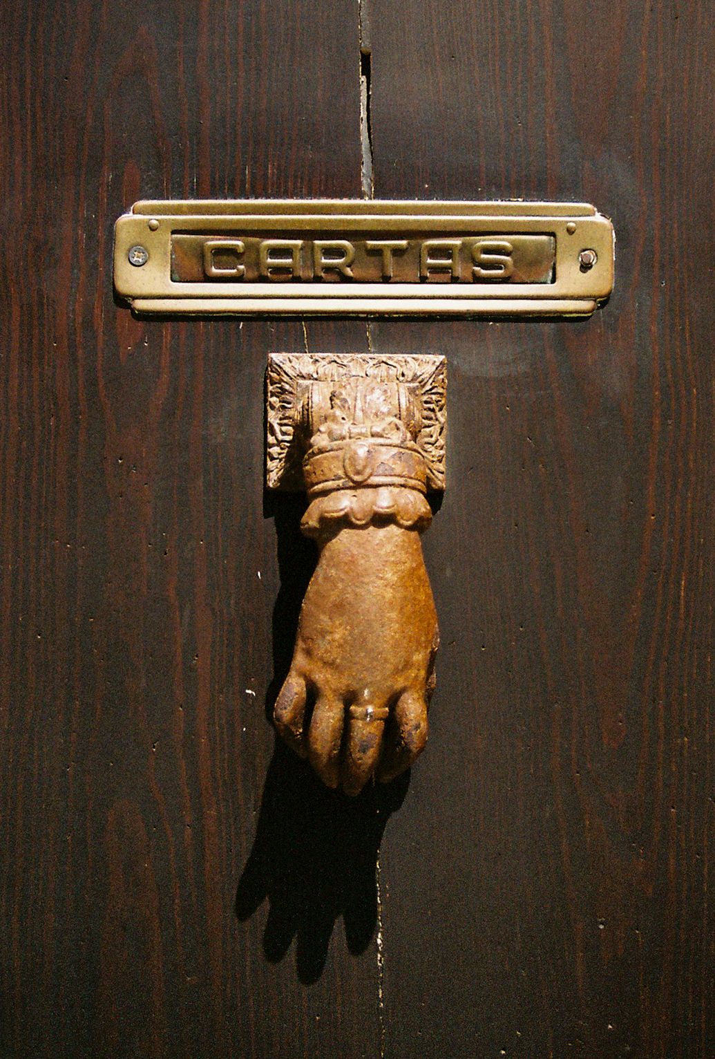 Ornate brass hand door knocker and cartas mail slot on a rustic dark wooden door.