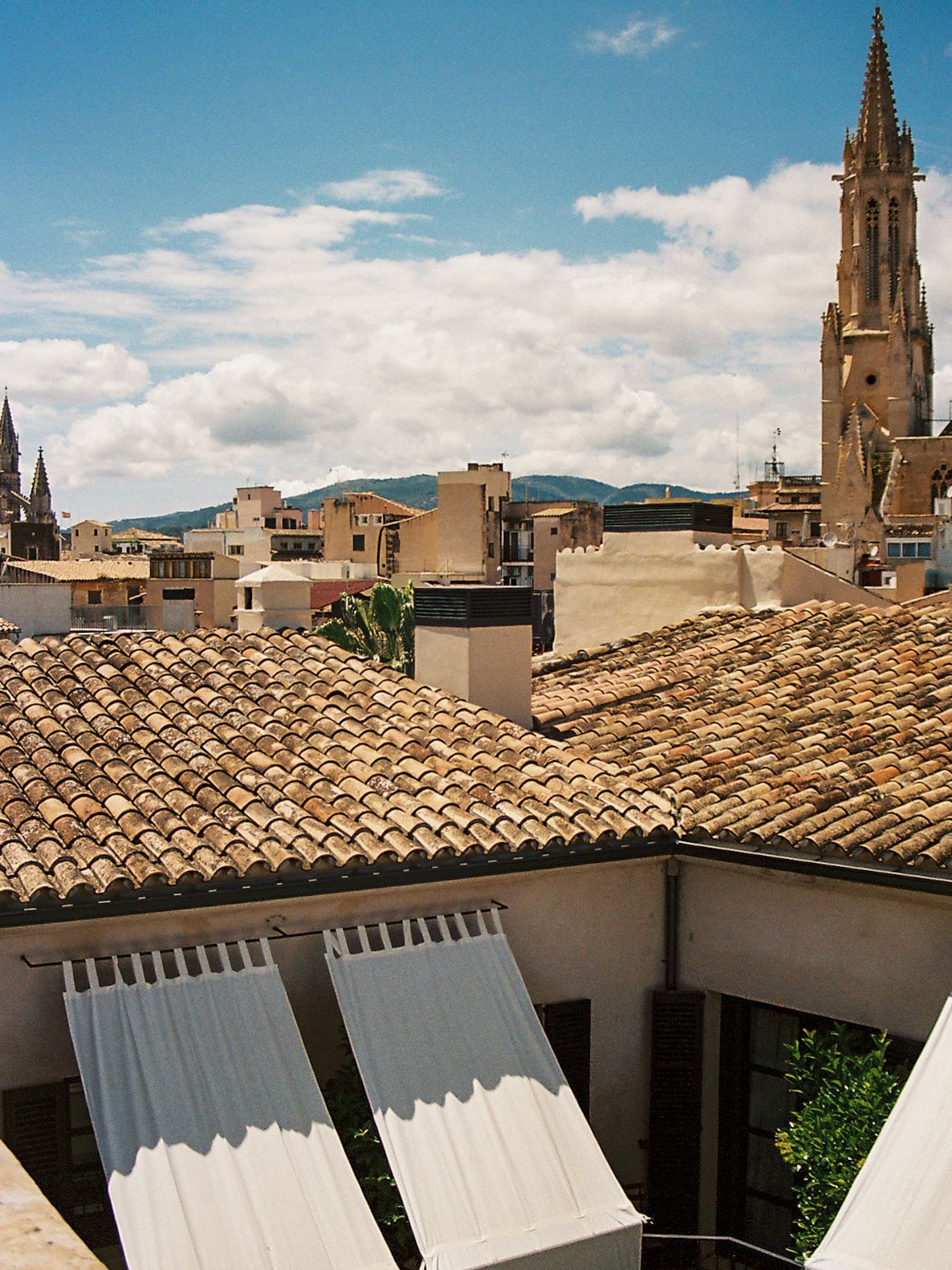 Rooftop view of terracotta tiled roofs with a Gothic church spire rising against a partly cloudy blue sky.