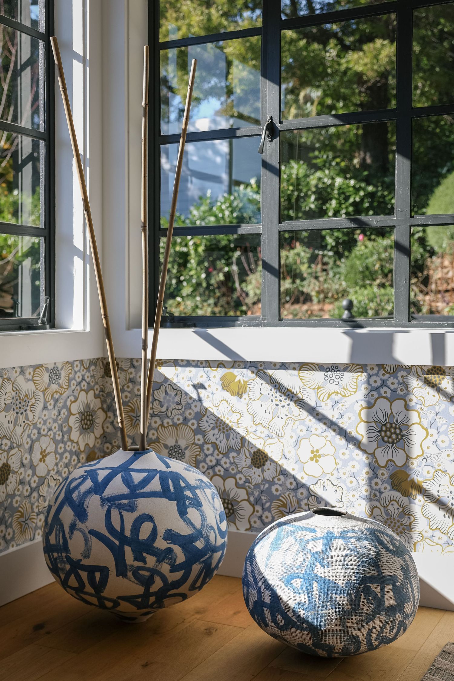Two blue and white ceramic vases with abstract patterns near a sunlit window with floral wallpaper.