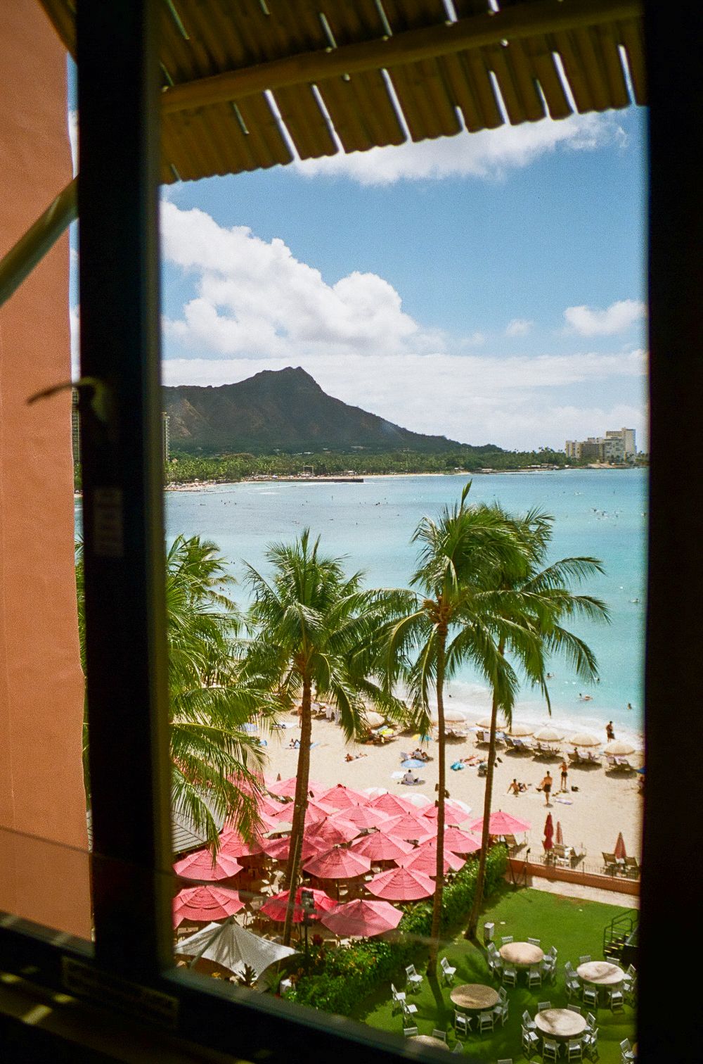 Tropical beach view through windows showing palm trees, red umbrellas, and Diamond Head mountain.