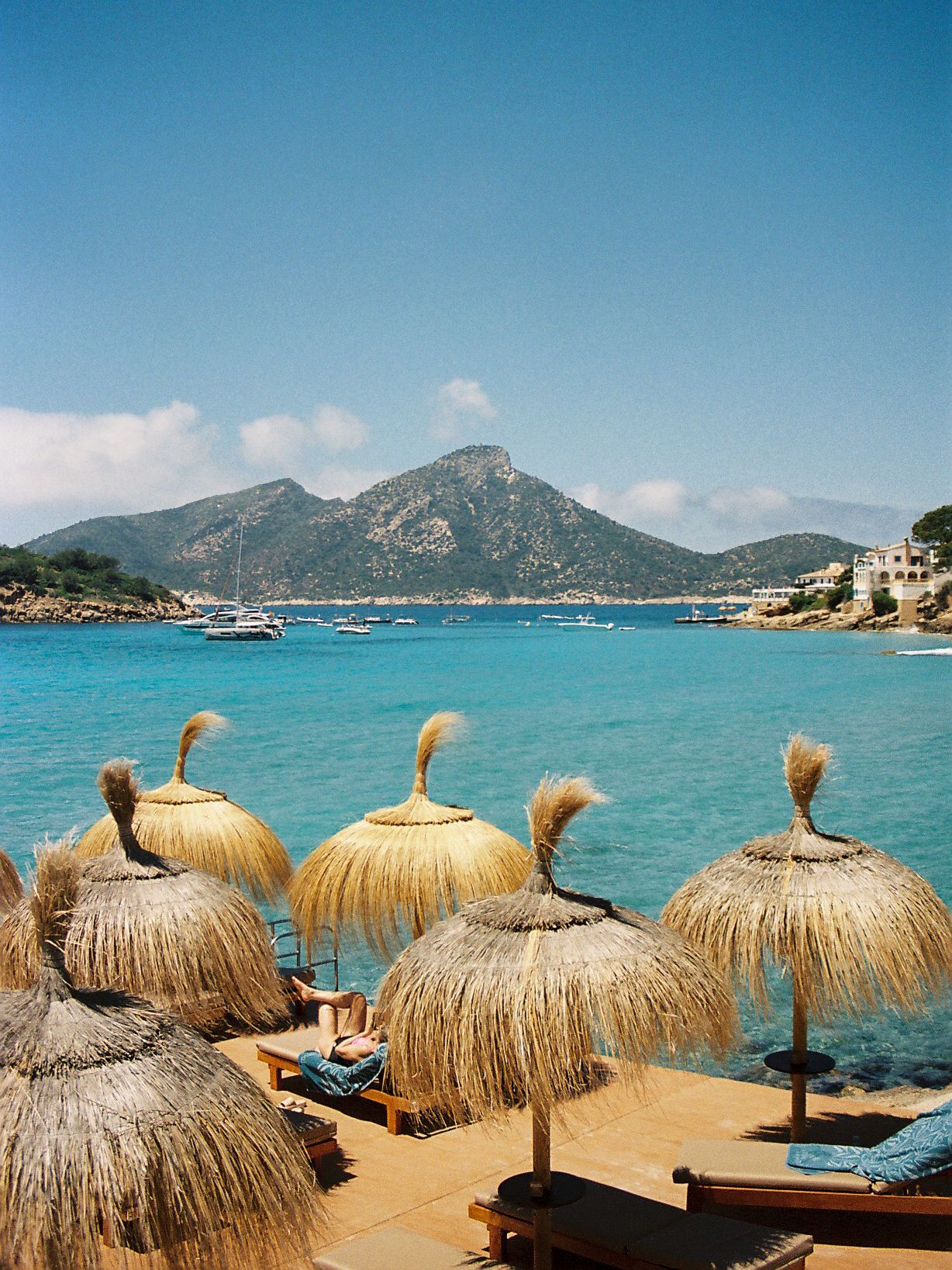 Thatched beach umbrellas overlook turquoise Mediterranean waters with moored boats and a mountain backdrop under clear blue skies.