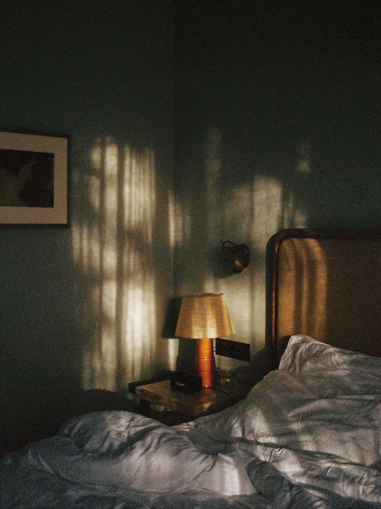 Moody bedroom with warm lamp light casting window shadows on dark green walls beside a wooden headboard.