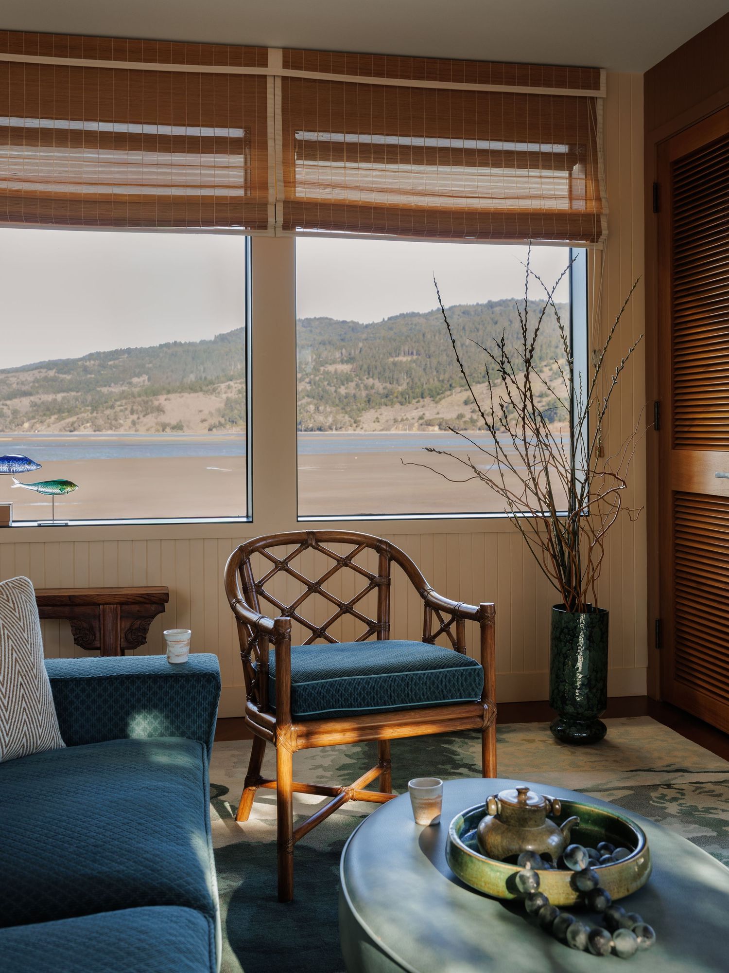 Cozy hotel lounge with rattan chairs, blue cushions, and panoramic windows overlooking a scenic lake and mountain view.