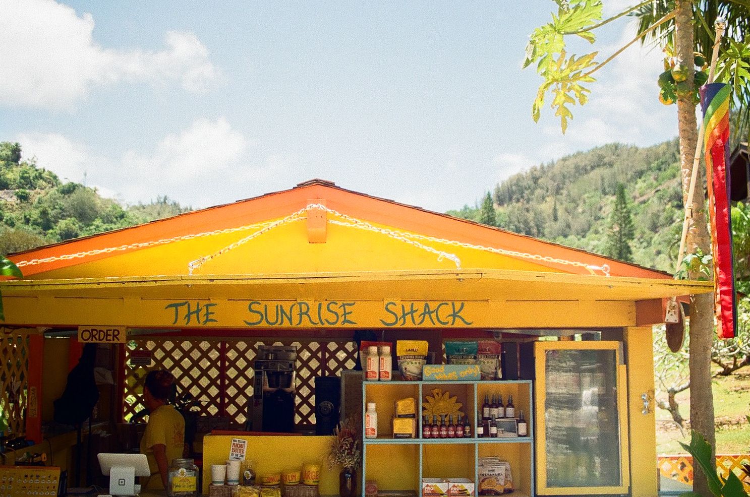 A bright yellow and orange food shack called 'The Sunrise Shack' with a lattice window display.