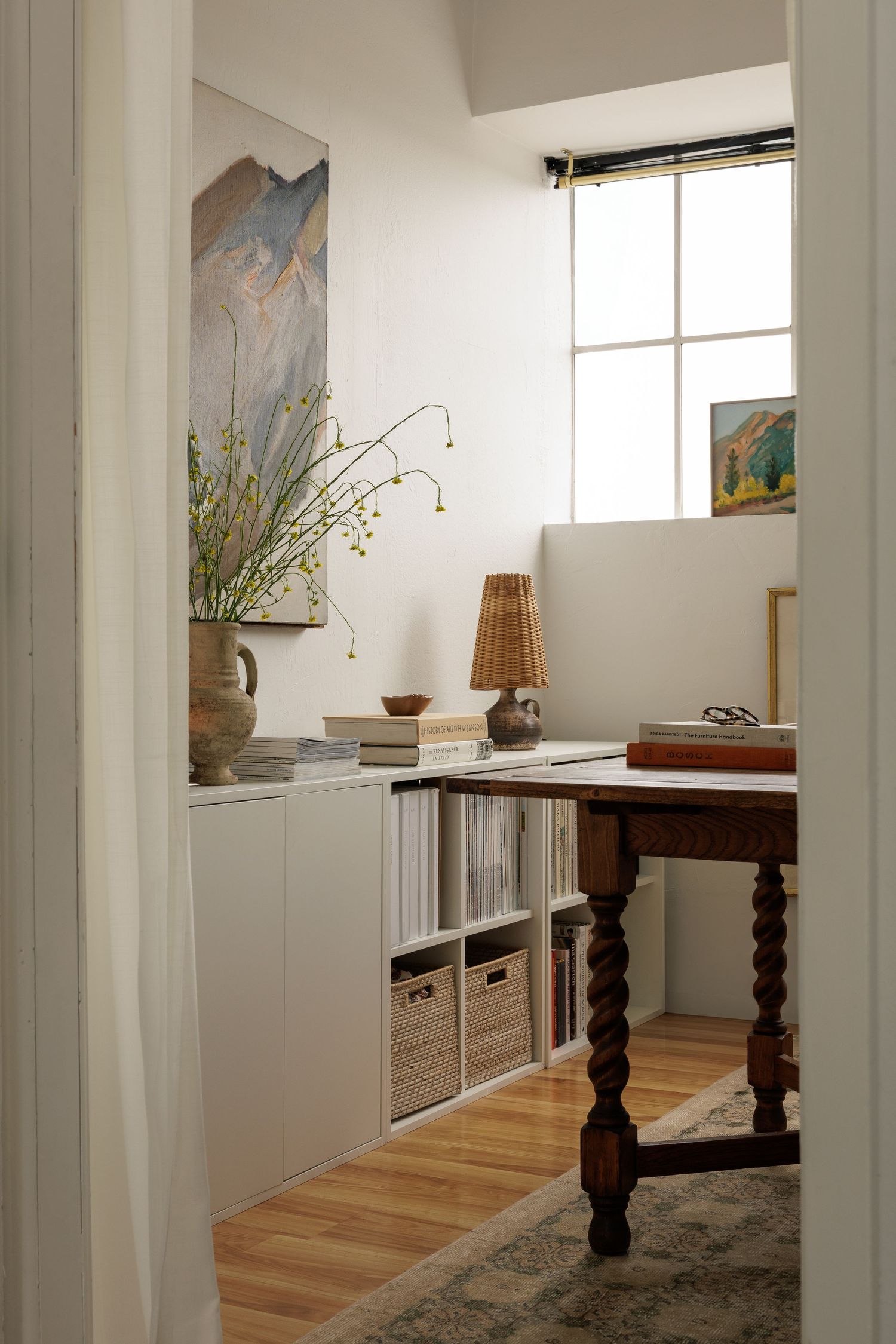Bright dining area with wooden table, natural light and artwork displayed on white walls.