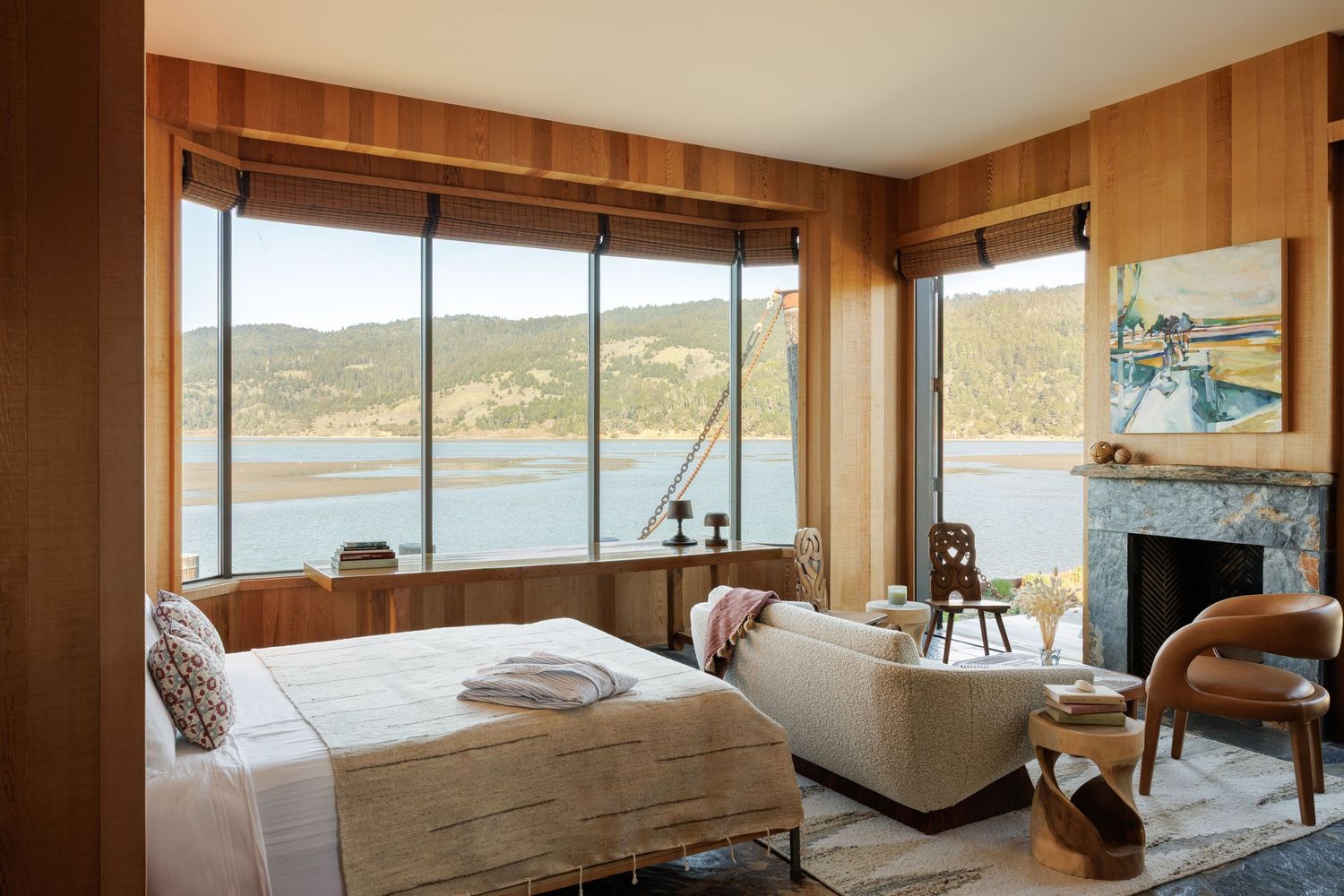 Cozy bedroom with wood-paneled walls, large windows overlooking a scenic lake and hills, a fireplace, and neutral furnishings.