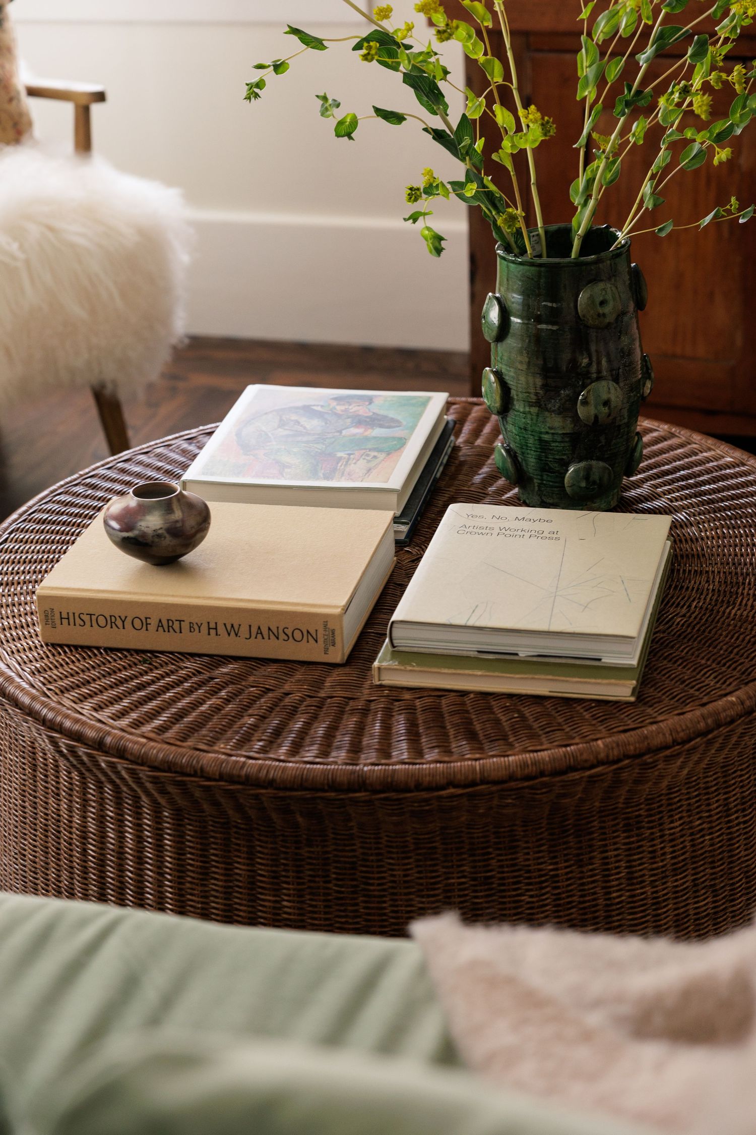 Rattan side table with stacked books and a green vase holding fresh greenery.