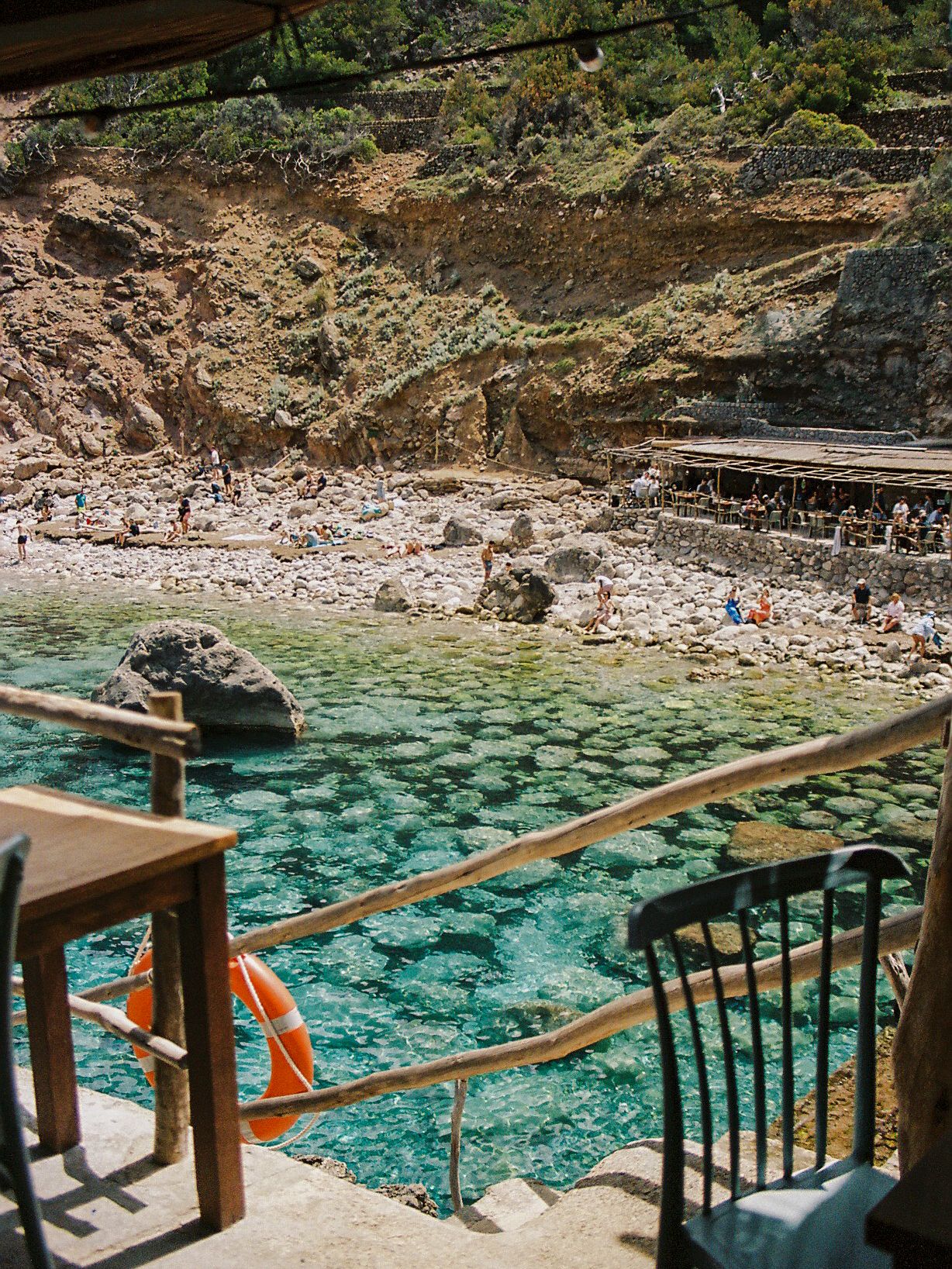 Crystal clear turquoise water cove with rocky cliffs, beach bar, and swimmers in a Mediterranean inlet.