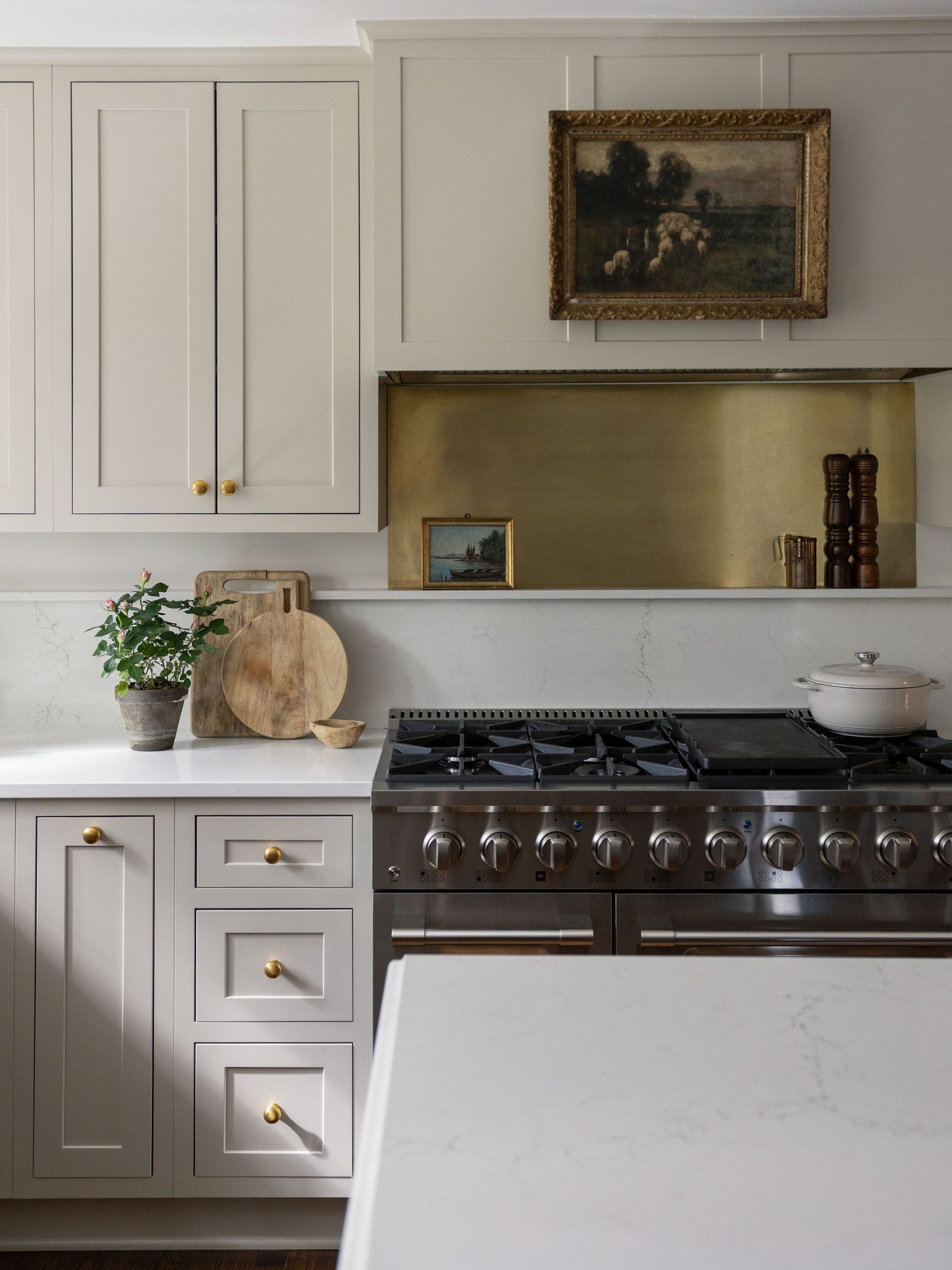 Elegant kitchen with greige shaker cabinets, brass hardware, marble countertops, dark range, and gold backsplash with vintage art.